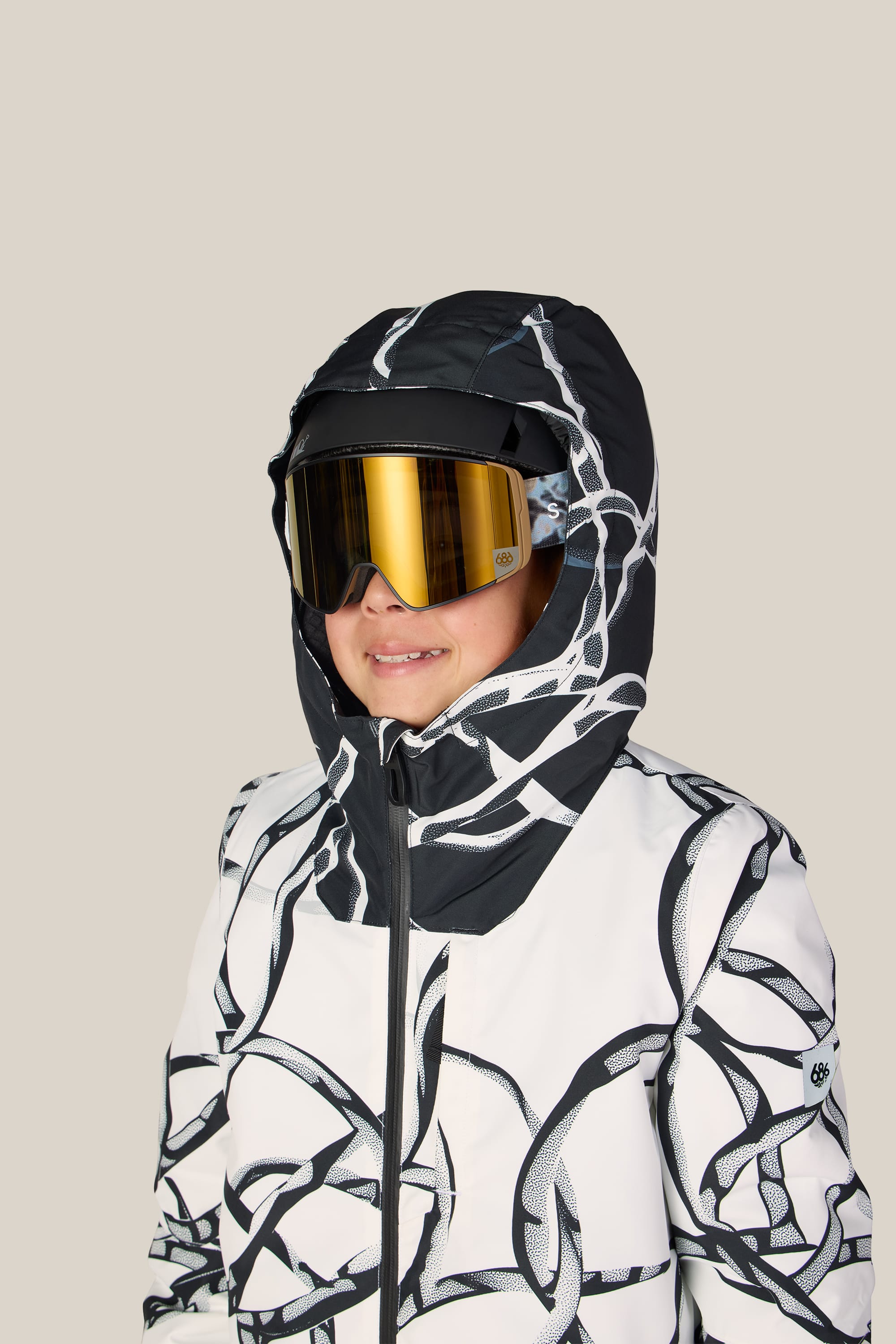 A child wearing the 686 Boys’ Gateway™ Insulated Jacket by 686, featuring black and white abstract patterns and reflective ski goggles, smiles slightly against a plain beige background.