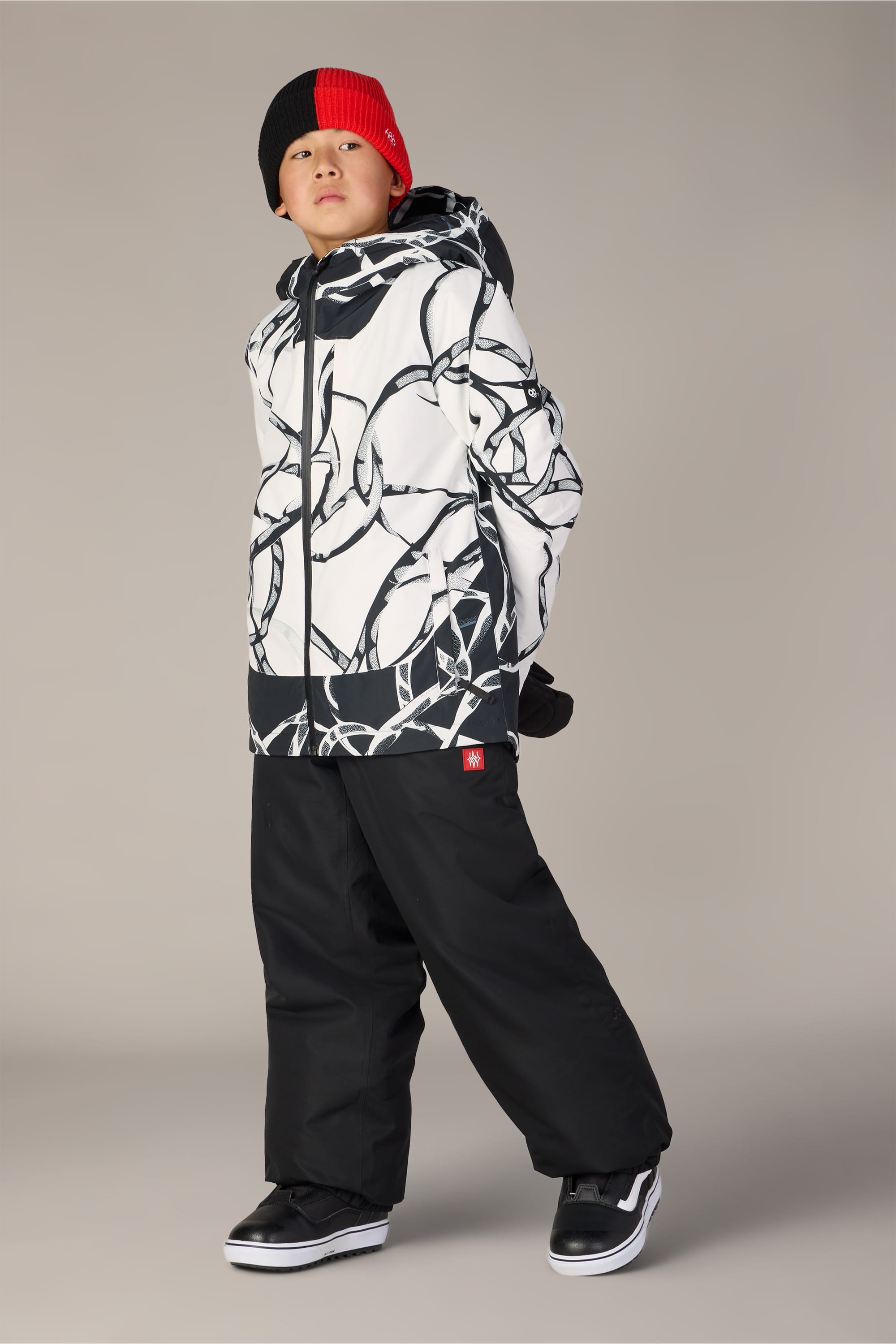A boy stands confidently against a plain background wearing the 686 Boys’ Gateway™ Insulated Jacket, featuring a white and black pattern, paired with black snow pants, black sneakers, black gloves, and a red and black beanie.