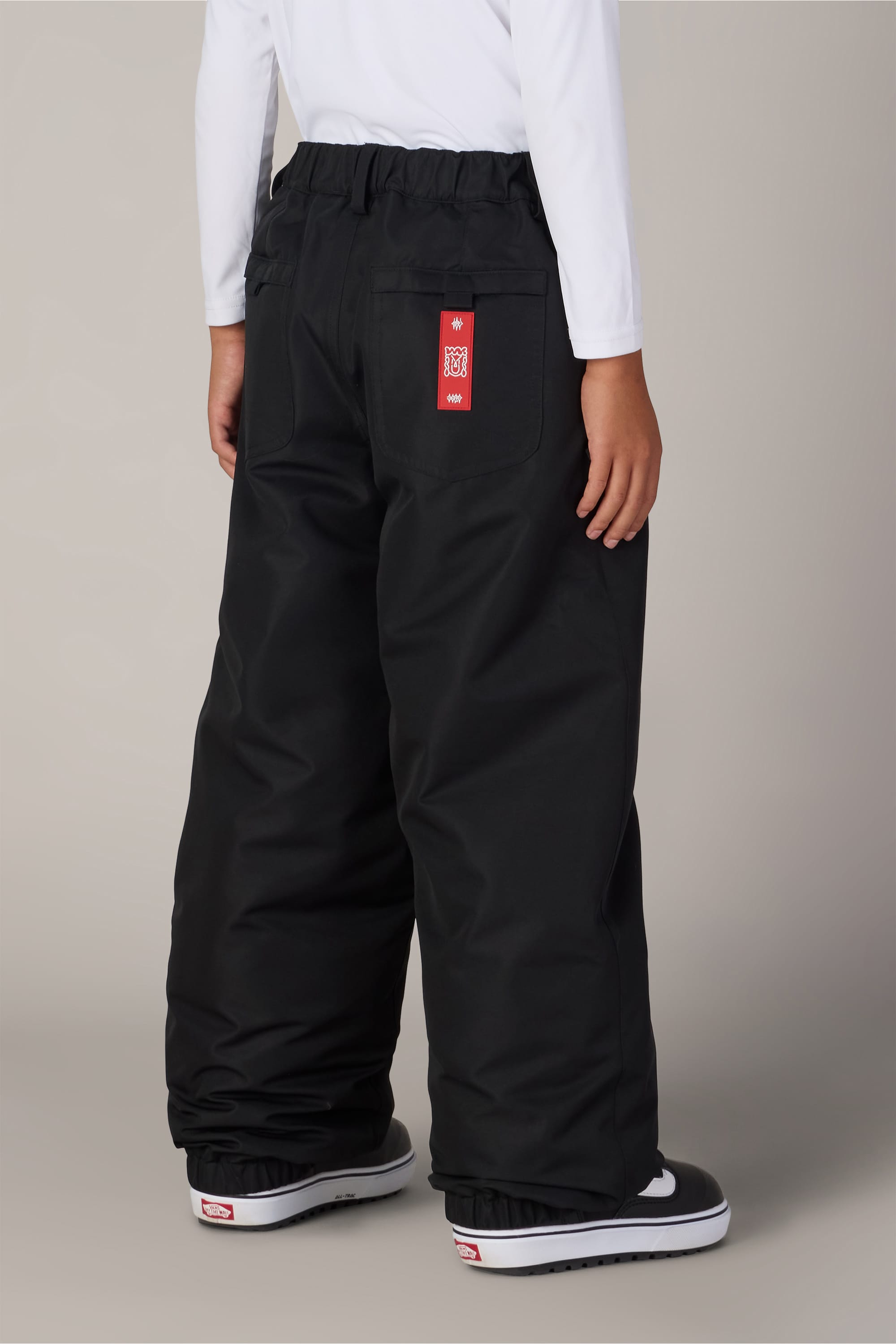 A child wearing the 686 Boys’ 686 Dojo® Insulated Pant in black, paired with a white long-sleeve shirt and black-and-white shoes, is shown from the back; a red tag is visible on the right back pocket of the pants.