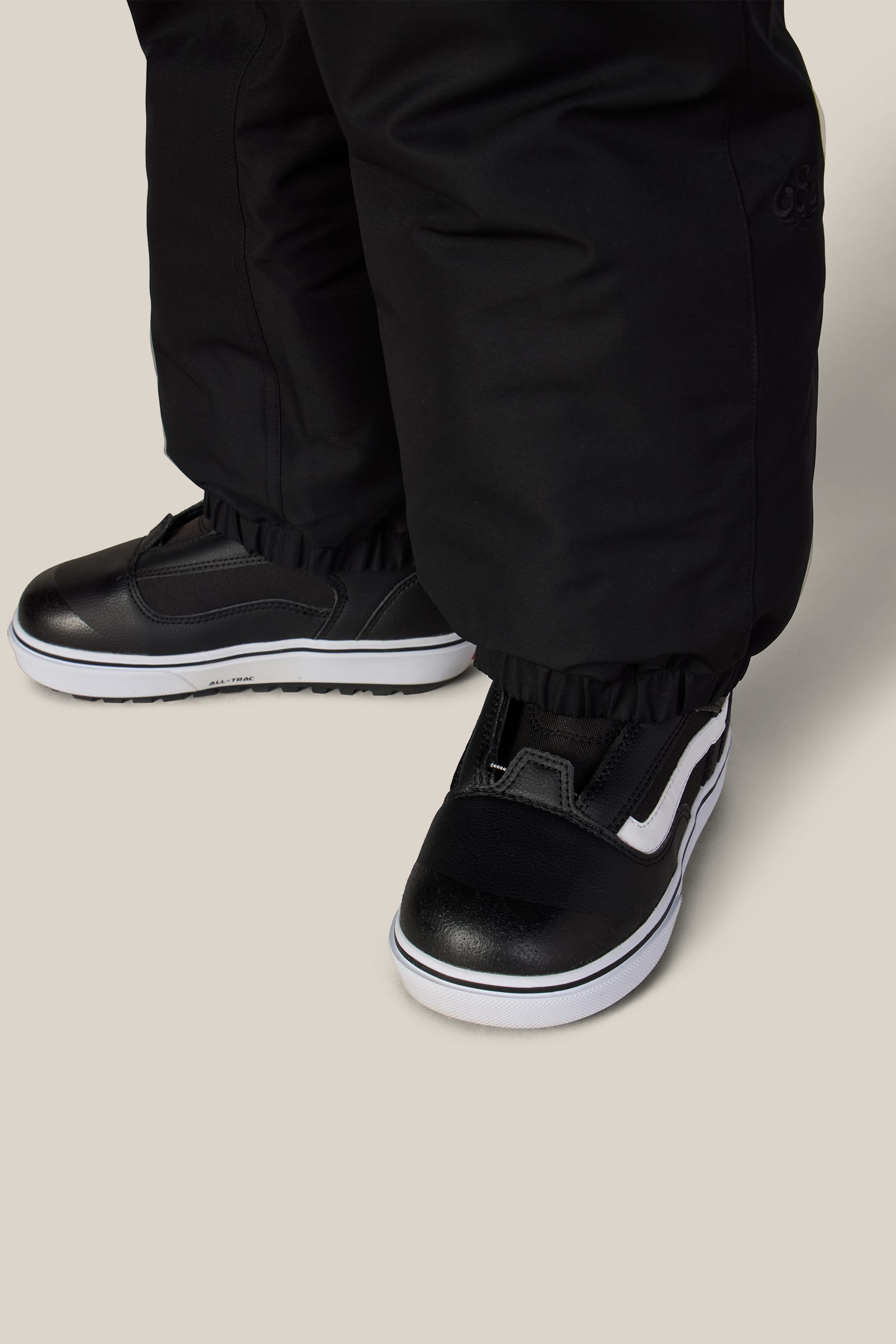 A close-up of a person wearing the 686 Boys’ 686 Dojo® Insulated Pant by 686, featuring black woven fabric with elastic cuffs, paired with black and white slip-on sneakers on a light beige background.