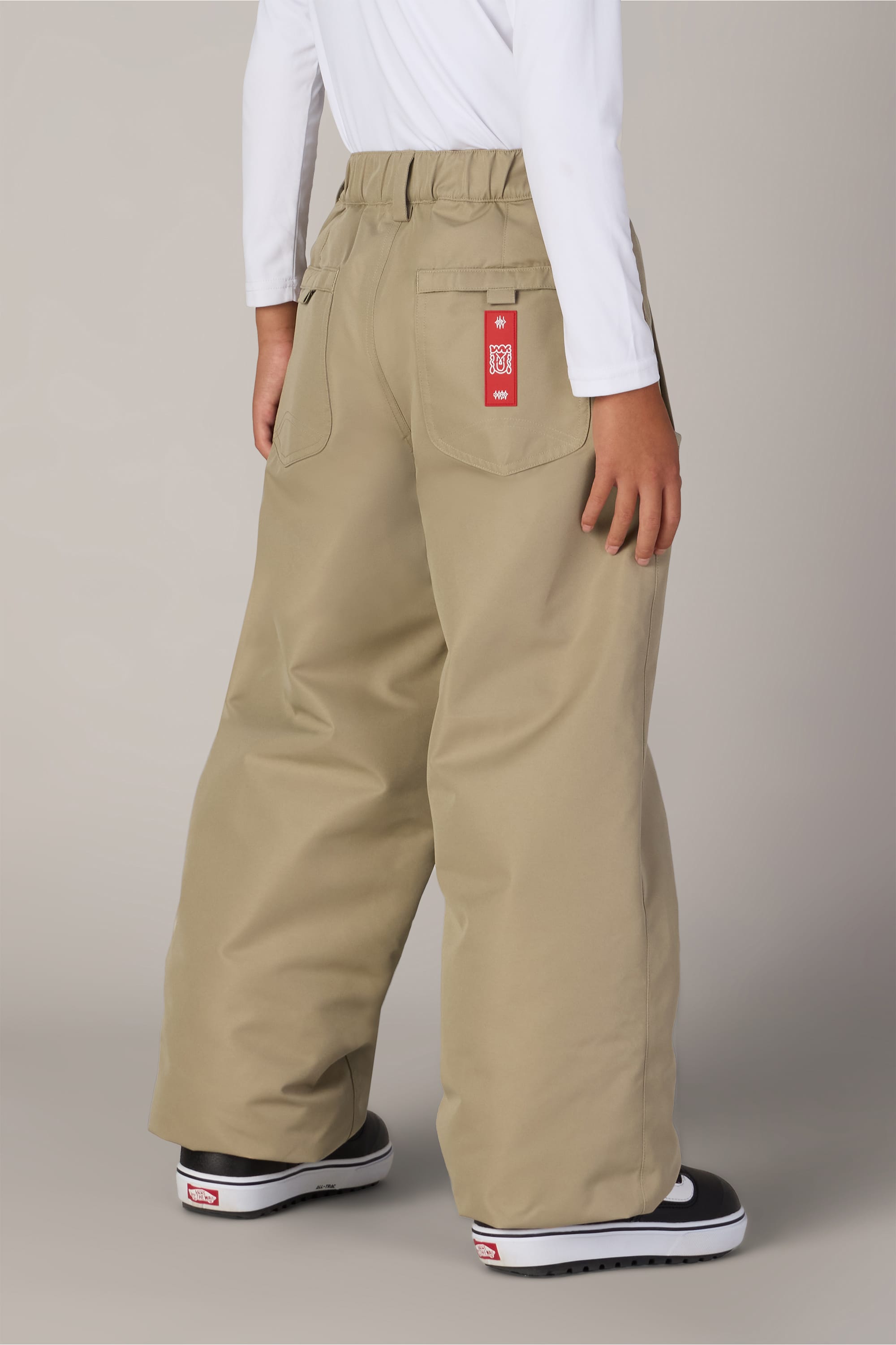 A child wearing 686 Boys’ 686 Dojo® Insulated Pant by 686 in a wide-leg khaki style with a red tag on the back pocket, paired with a white long-sleeve shirt and black Vans sneakers, stands facing away from the camera.