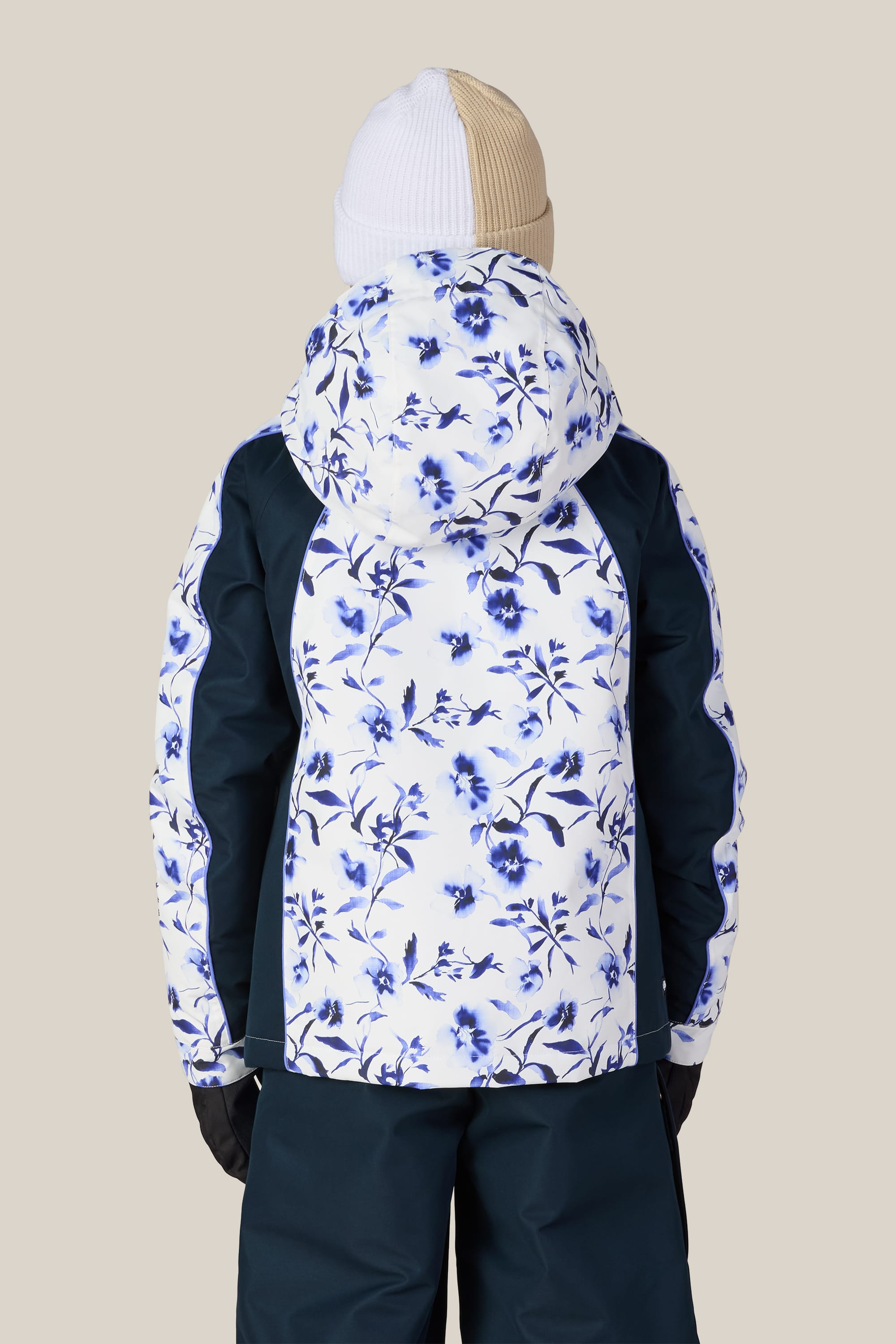 A person facing away is wearing the 686 Girls’ Hologram™ Insulated Jacket by 686, which features a white and blue floral pattern with dark sleeves. They are also dressed in dark pants, black gloves, and a two-tone knit hat, set against a plain beige background.