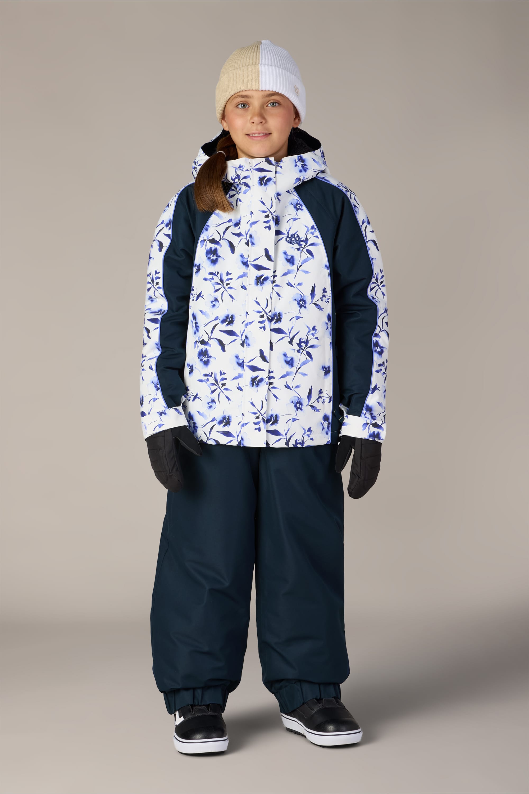 A smiling child stands in winter gear against a plain beige background. She wears the 686 Girls’ Hologram™ Insulated Jacket—a blue and white floral-patterned waterproof jacket from 686—paired with dark snow pants, black gloves, black boots, and a white-beige beanie. Her long brown hair cascades beneath her hat.