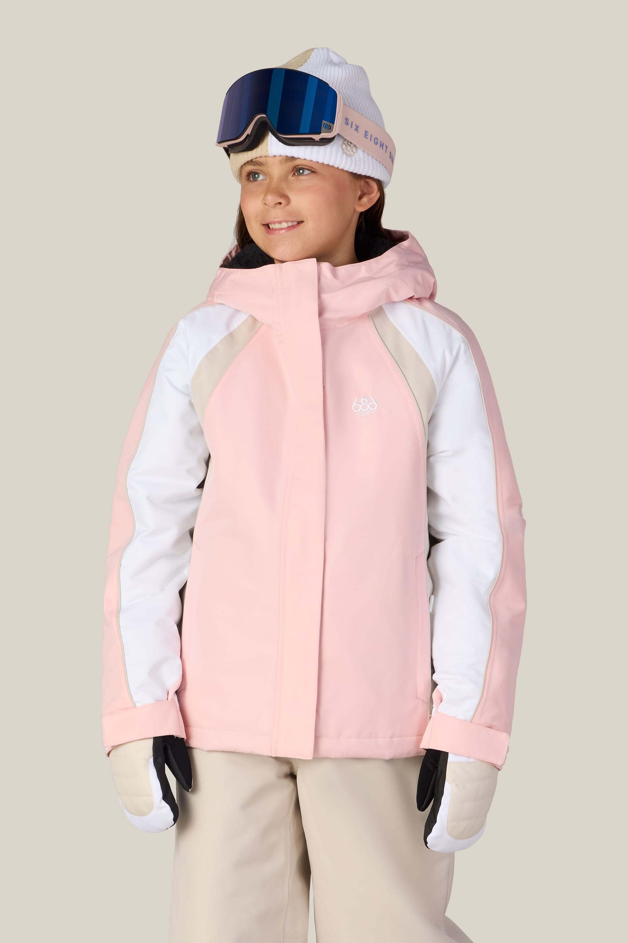A girl wearing the 686 Girls’ Hologram™ Insulated Jacket by 686, paired with white gloves, beige pants, a white beanie, and blue ski goggles stands smiling against a plain background.