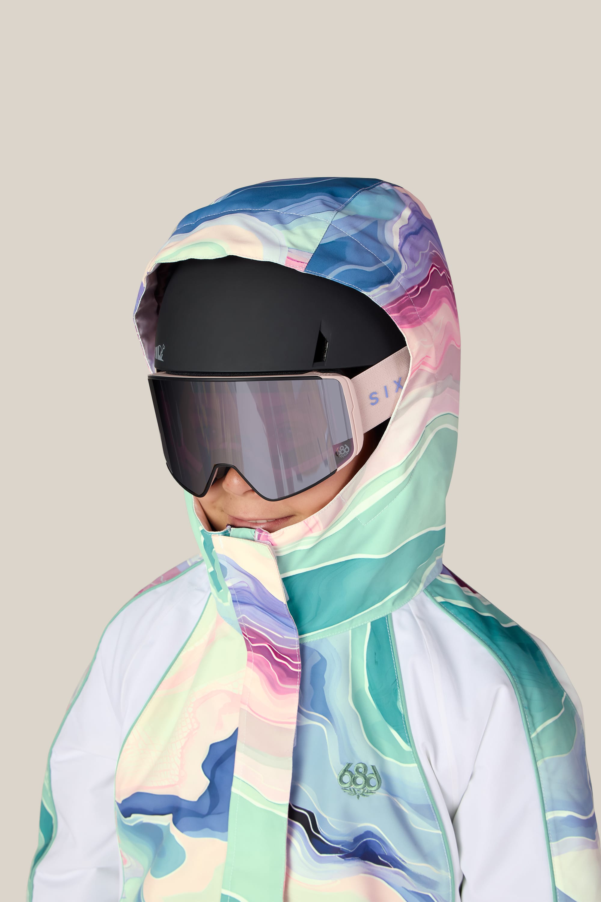 A child wearing the 686 Girls’ Hologram™ Insulated Jacket—a pastel-patterned, waterproof hooded jacket by 686—paired with ski goggles and a black helmet, facing slightly left against a plain beige background.