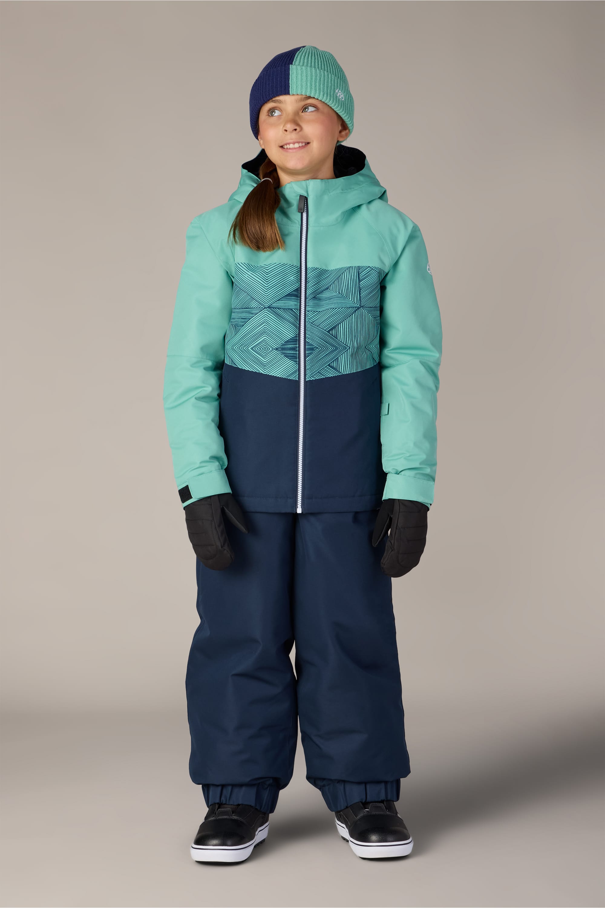 A young girl wearing a teal and navy winter jacket, 686 Girls’ Outline™ Insulated Pant by 686 in a matching navy shade, black gloves, a color-block beanie, and black shoes stands smiling against a plain gray background.