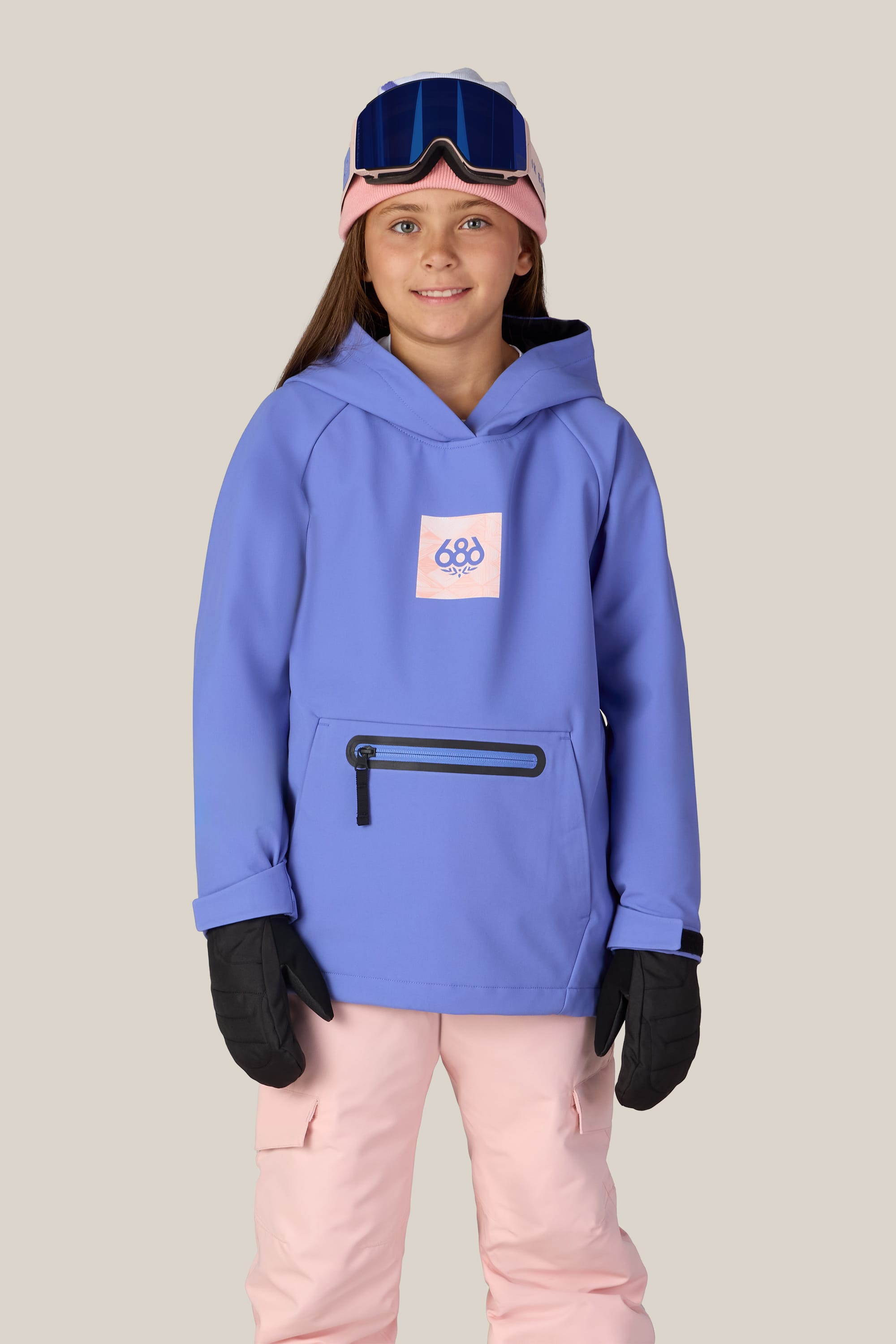 A smiling child wearing the 686 Youth Waterproof Hoody ski jacket in blue, featuring a pink 686 logo, paired with pink snow pants, black gloves, ski goggles, and a pink headband stands against a plain light background.