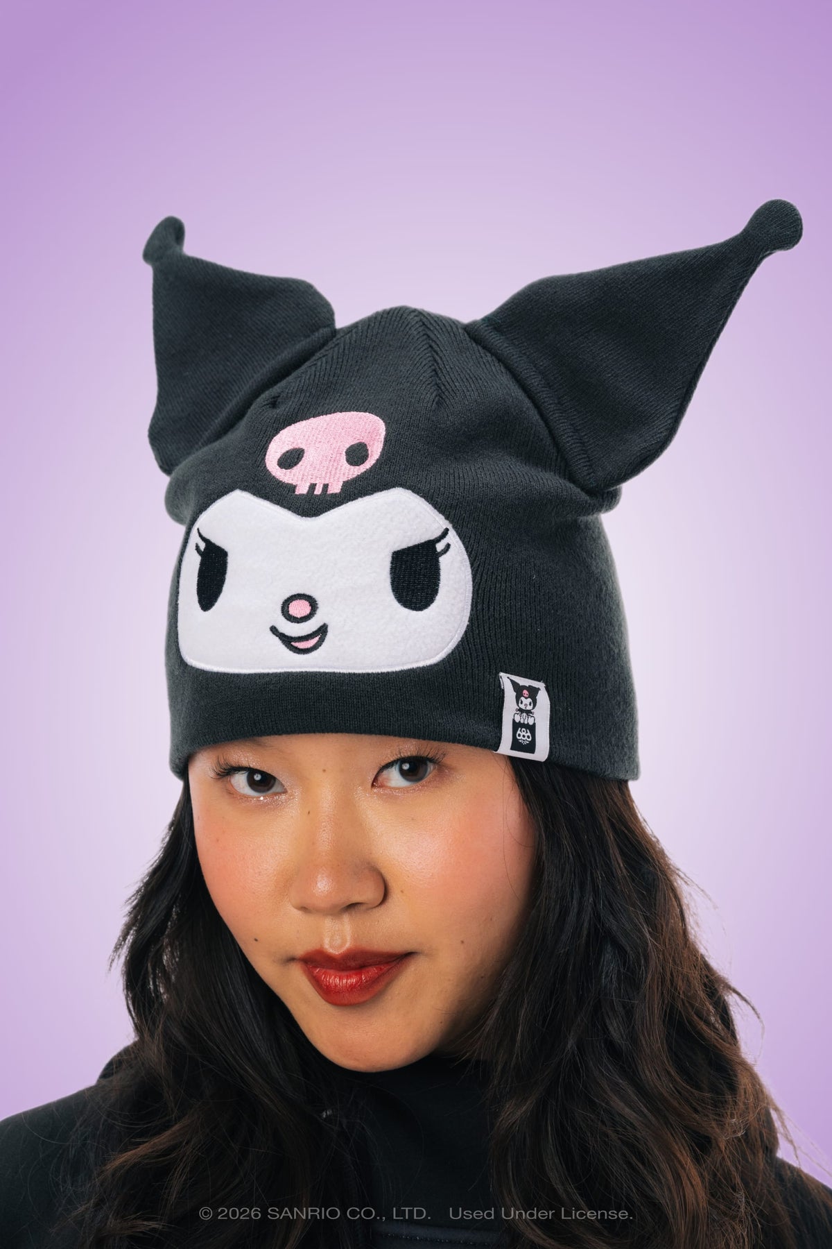 A person wearing the 686 Women’s Kuromi™ Beanie by 686, featuring a black acrylic knit design with pointed ears, a white face, and a pink skull detail, set against a purple gradient background.