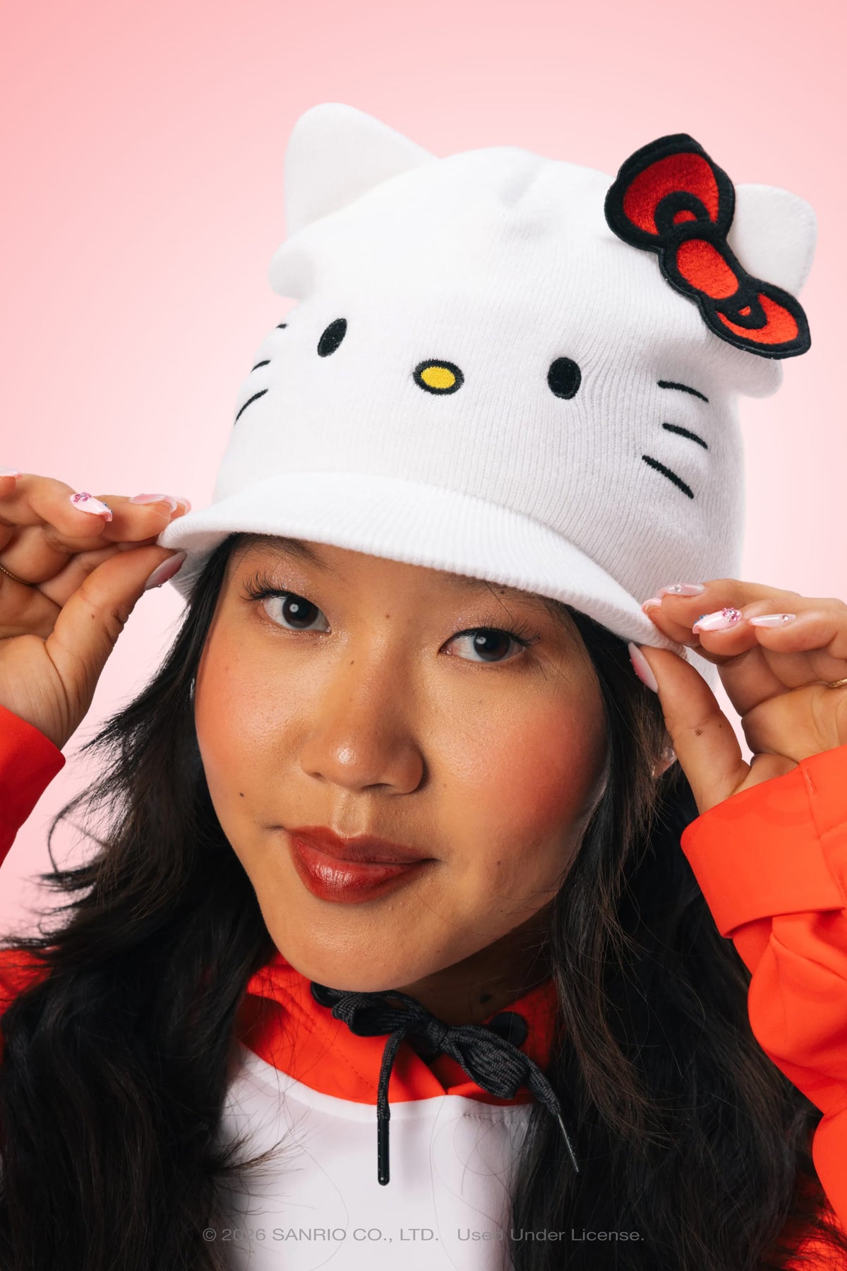 A woman wearing the 686 Women’s Hello Kitty® Visor Beanie, featuring a red bow, gently holds the brim and smiles softly against a pink background.