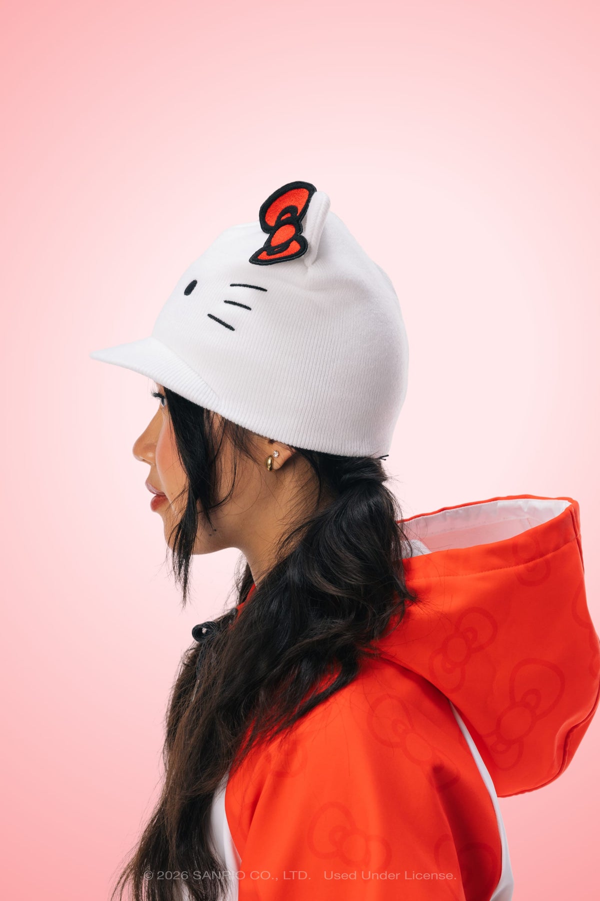 A person in profile wears the 686 Women’s Hello Kitty® Visor Beanie—a white acrylic knit beanie with Hello Kitty ears and a red bow—from 686, along with an orange jacket featuring Hello Kitty patterns, set against a pink background.