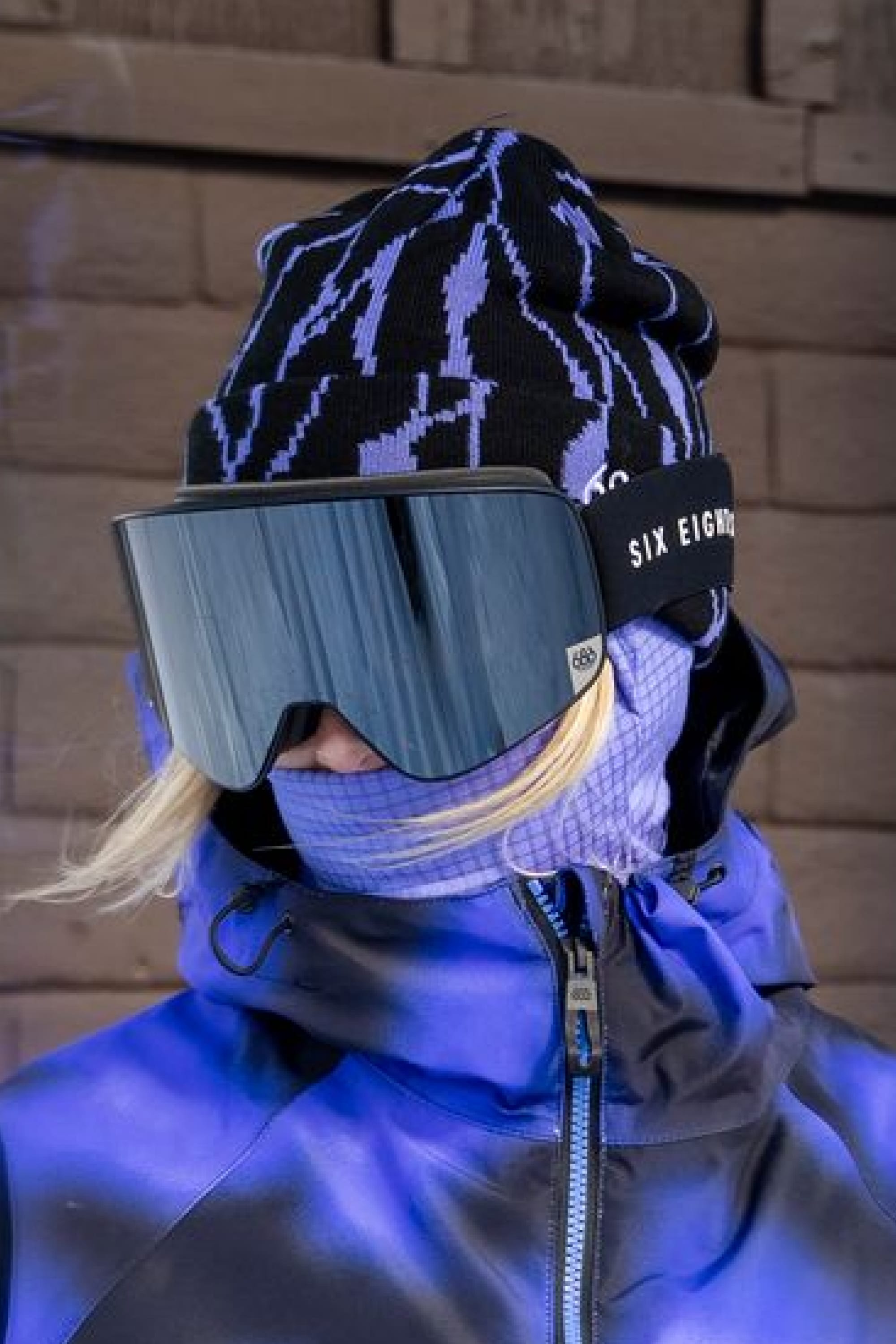 A person in a blue and purple winter jacket, wearing the 686 Women’s Cepa Beanie by 686, ski goggles, and a face covering, standing in front of a wooden wall.