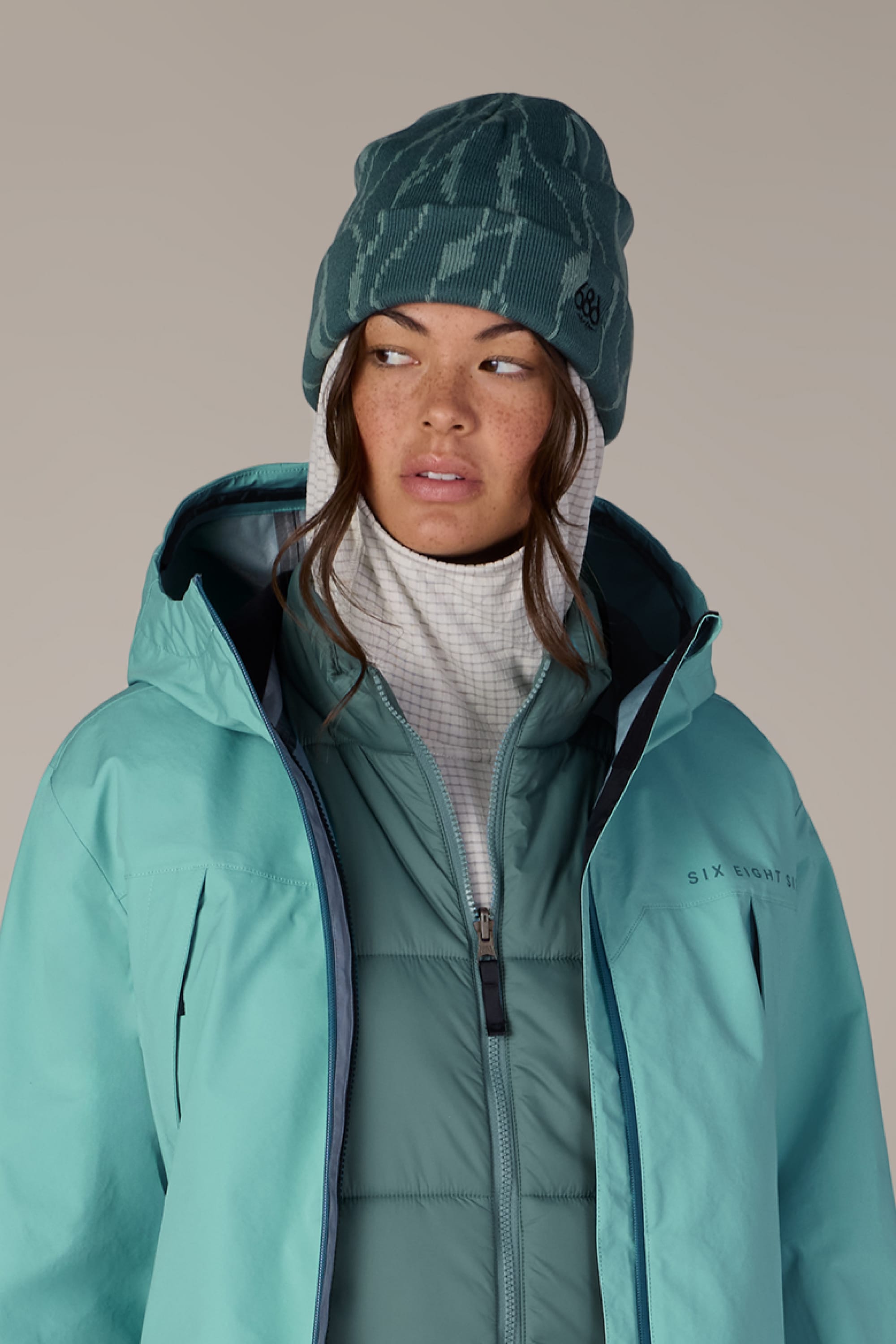 Wearing the 686 Women’s Cepa Beanie, a person in teal winter clothing looks to the side. They pair the beanie with a hooded jacket, a puffer vest, and a high-collared white base layer, set against a plain background.