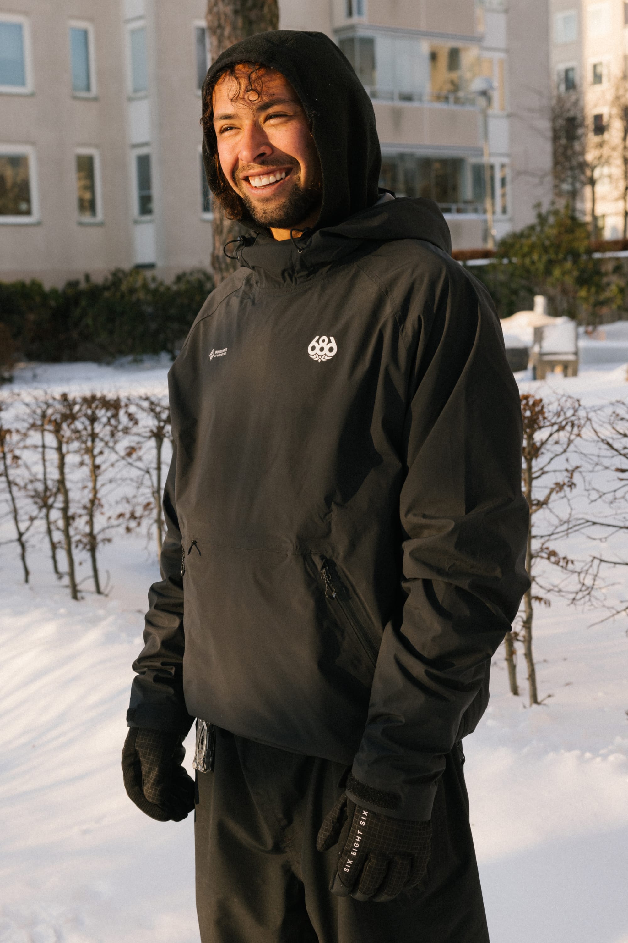 A person wearing the 686 Men’s WINDSTOPPER® Pullover Hoody by GORE-TEX® LABS stands outside in a snowy area, smiling, with apartment buildings and bare bushes in the background.