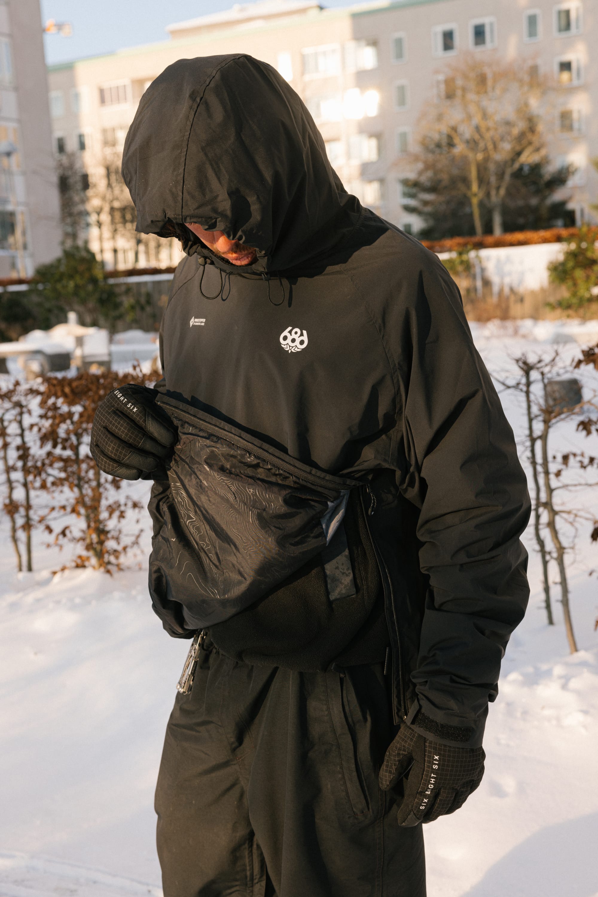 A person wearing the 686 Men’s WINDSTOPPER® Pullover Hoody by GORE-TEX® LABS stands outside in the snow, looking into a black crossbody bag. Apartment buildings and bare bushes are visible in the background.