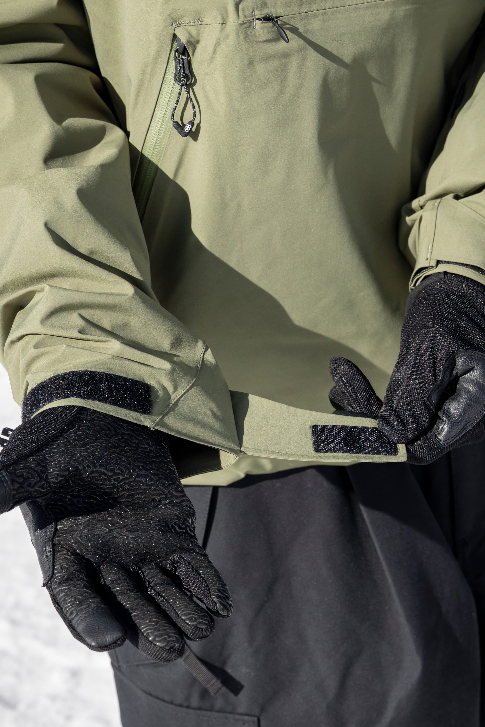 A person wearing black gloves and the 686 Men’s WINDSTOPPER® Pullover Hoody by GORE-TEX® LABS adjusts the velcro strap on their sleeve, with snow visible in the background.