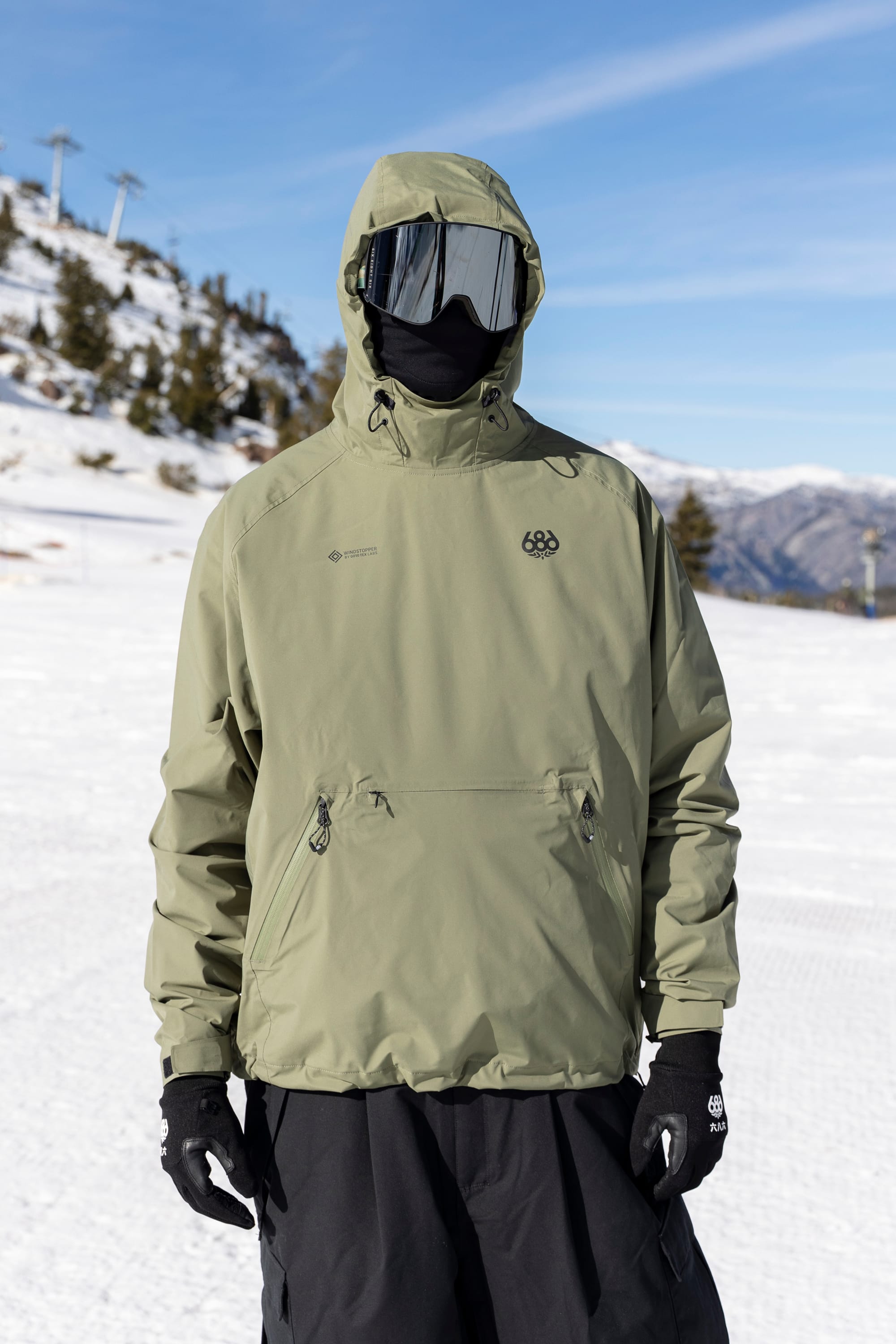 A person wearing the 686 Men’s WINDSTOPPER® Pullover Hoody by GORE-TEX® LABS, along with black gloves, black pants, and a reflective ski mask, stands on a snowy mountain surrounded by trees and ski lifts under a clear blue sky.