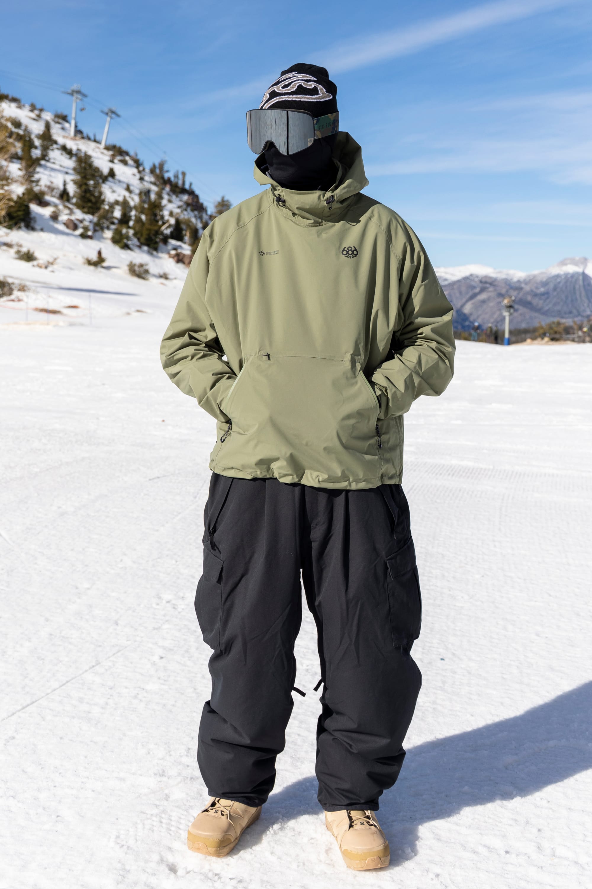 A person stands on a snowy slope wearing the 686 Men’s WINDSTOPPER® Pullover Hoody by GORE-TEX® LABS in green, black snow pants, tan boots, black gloves, a black hat, and large ski goggles, with mountains and trees in the background beneath a blue sky.