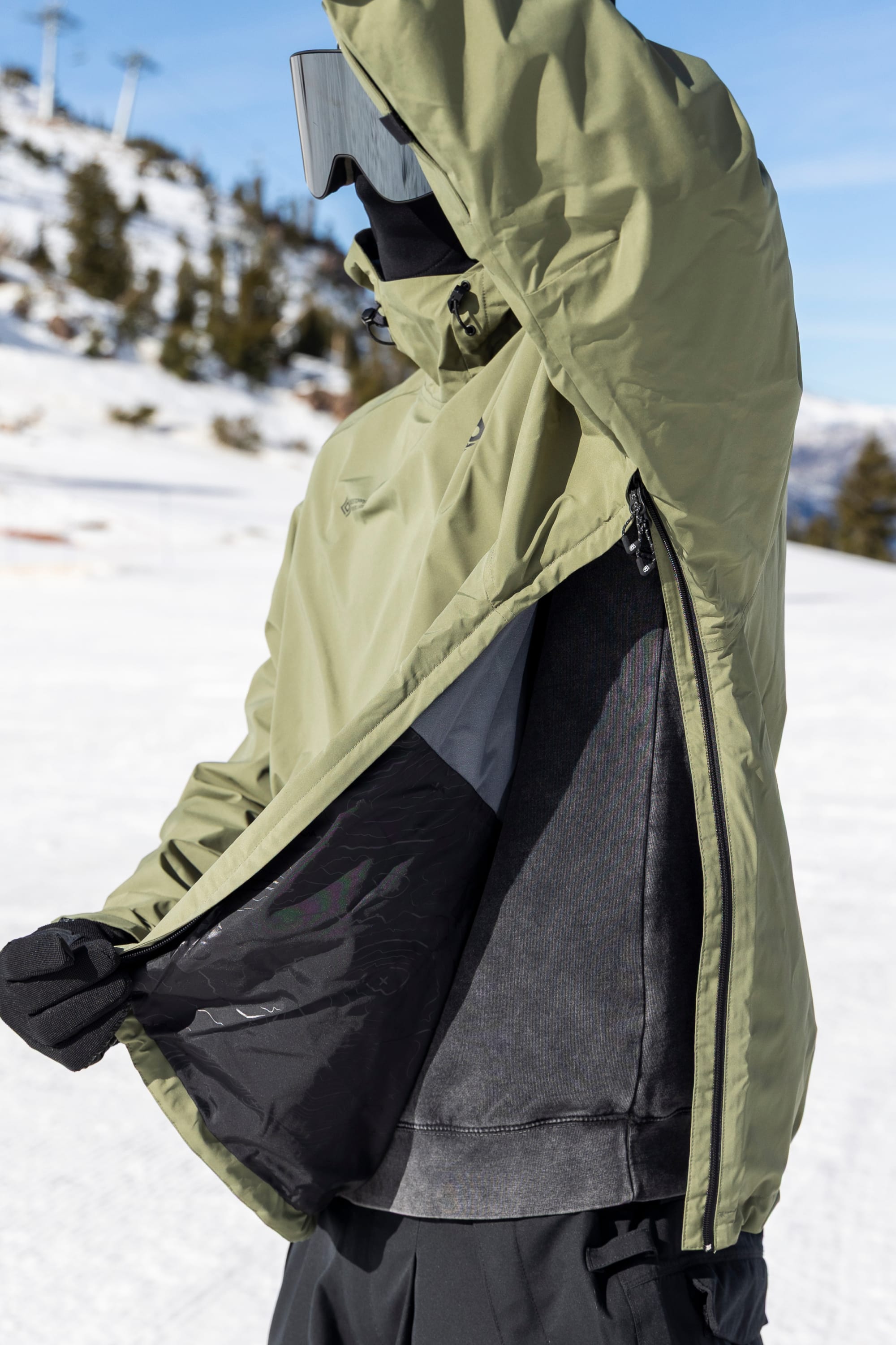 A person wearing the 686 Men’s WINDSTOPPER® Pullover Hoody by GORE-TEX® LABS in green opens a side zipper to reveal inner layers while standing on a snowy mountain, with trees and ski lifts visible in the background.