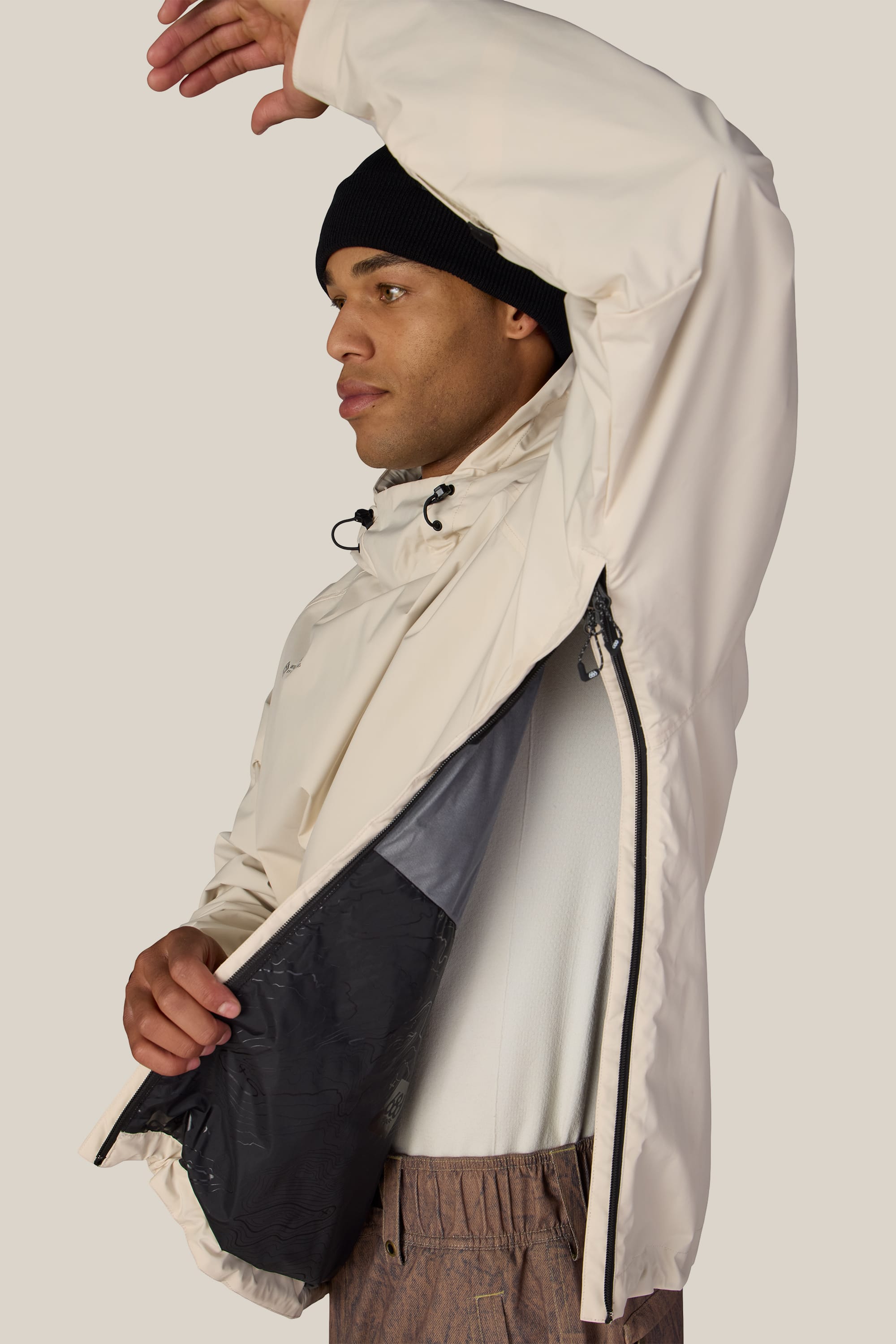 Wearing a 686 Men’s WINDSTOPPER® Pullover Hoody by GORE-TEX® LABS and a black beanie, a person lifts their arm to reveal the inner lining and convenient ventilation zipper of the cream-colored hoody.