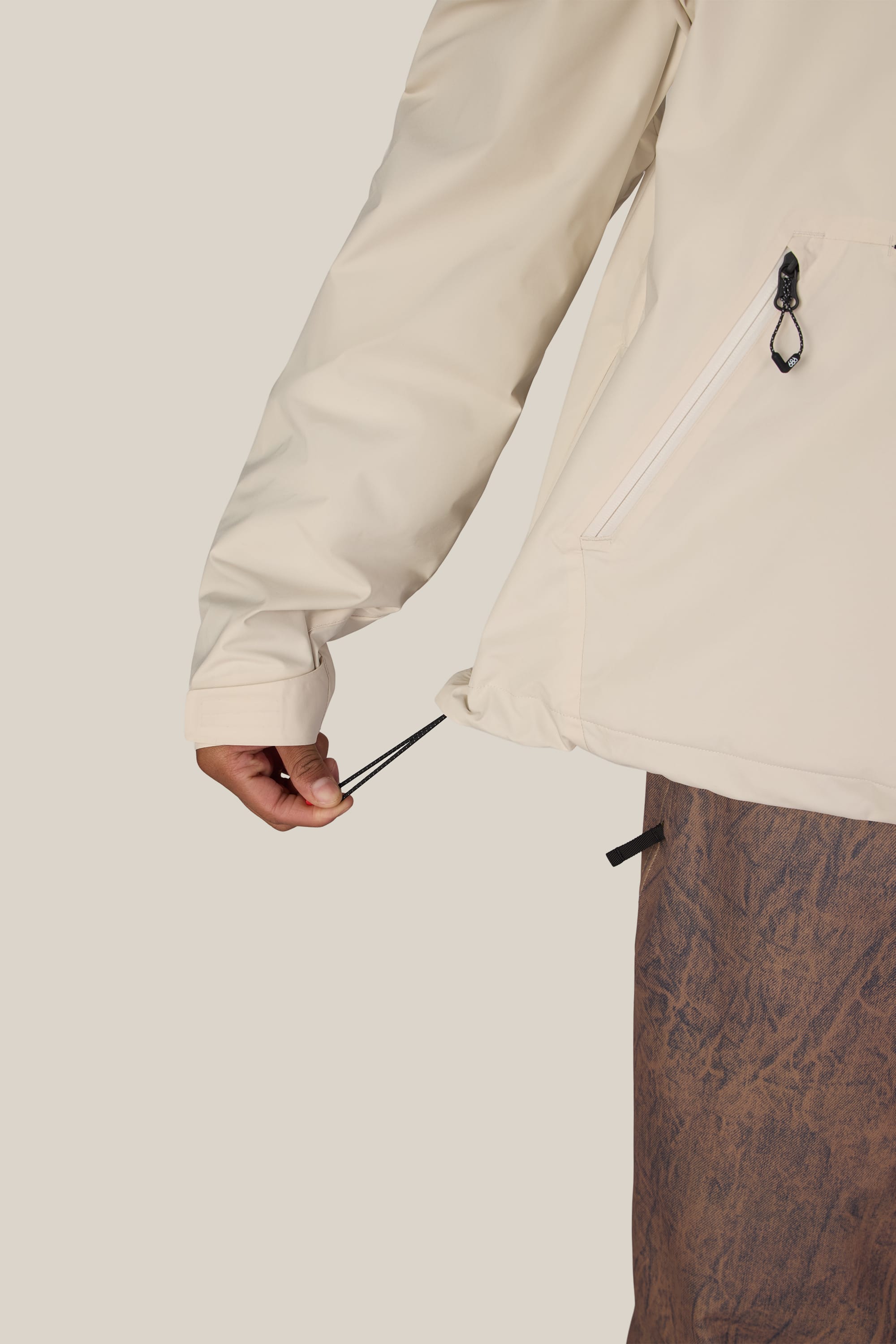 A person adjusts the drawstring of the 686 Men’s WINDSTOPPER® Pullover Hoody by GORE-TEX® LABS in light beige, paired with brown textured pants, against a plain background.