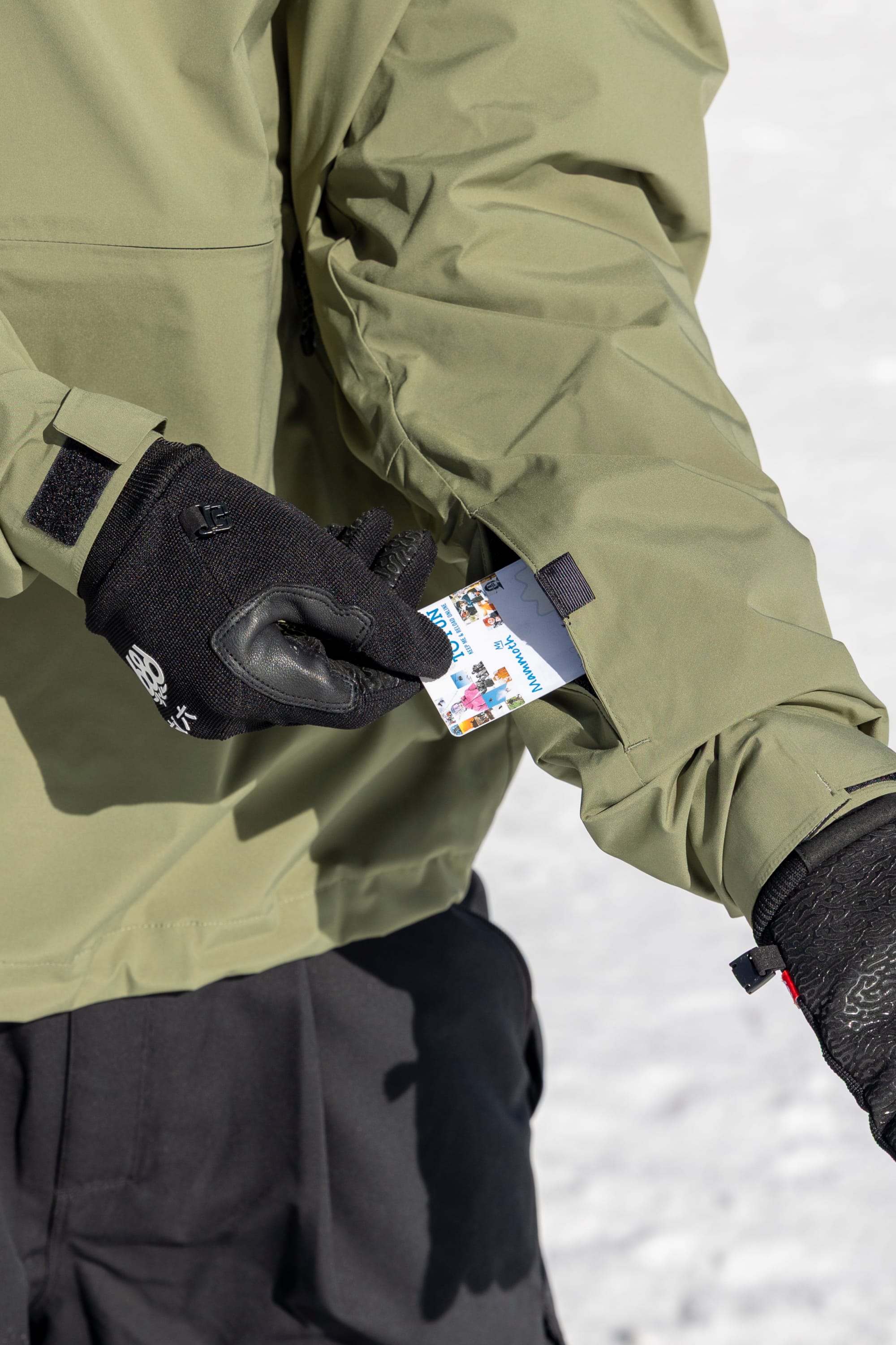Wearing black gloves and the 686 Men’s WINDSTOPPER® Pullover Hoody by GORE-TEX® LABS in olive green, a person pulls a ski pass from a sleeve pocket with snow visible in the background.