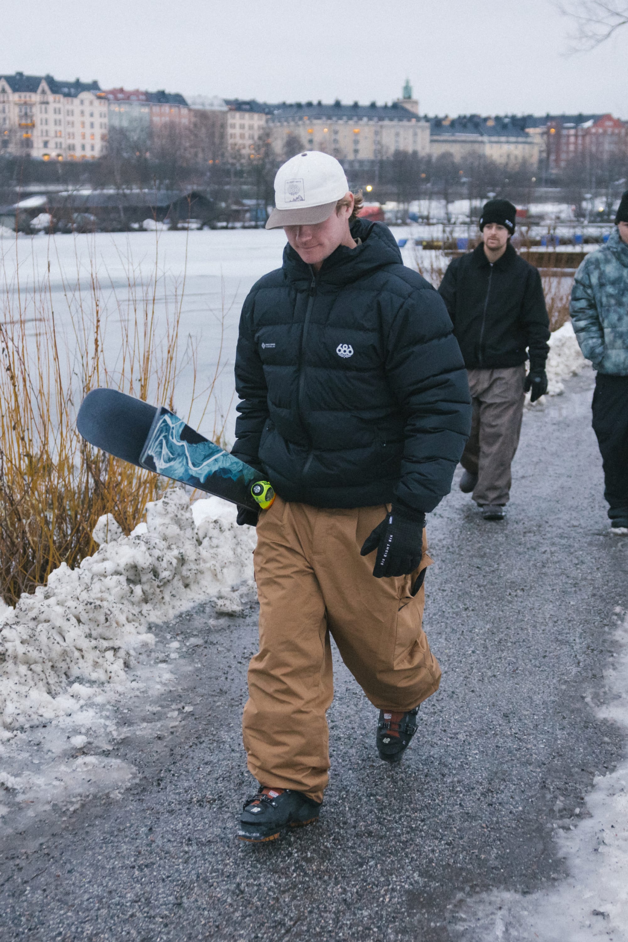 A person wearing the 686 Men’s WINDSTOPPER® Pyro™ Down Puffer Jacket by GORE-TEX® LABS, along with tan pants and a white cap, walks on a snowy path holding a snowboard. Two others follow behind, with buildings and a frozen river in the background.