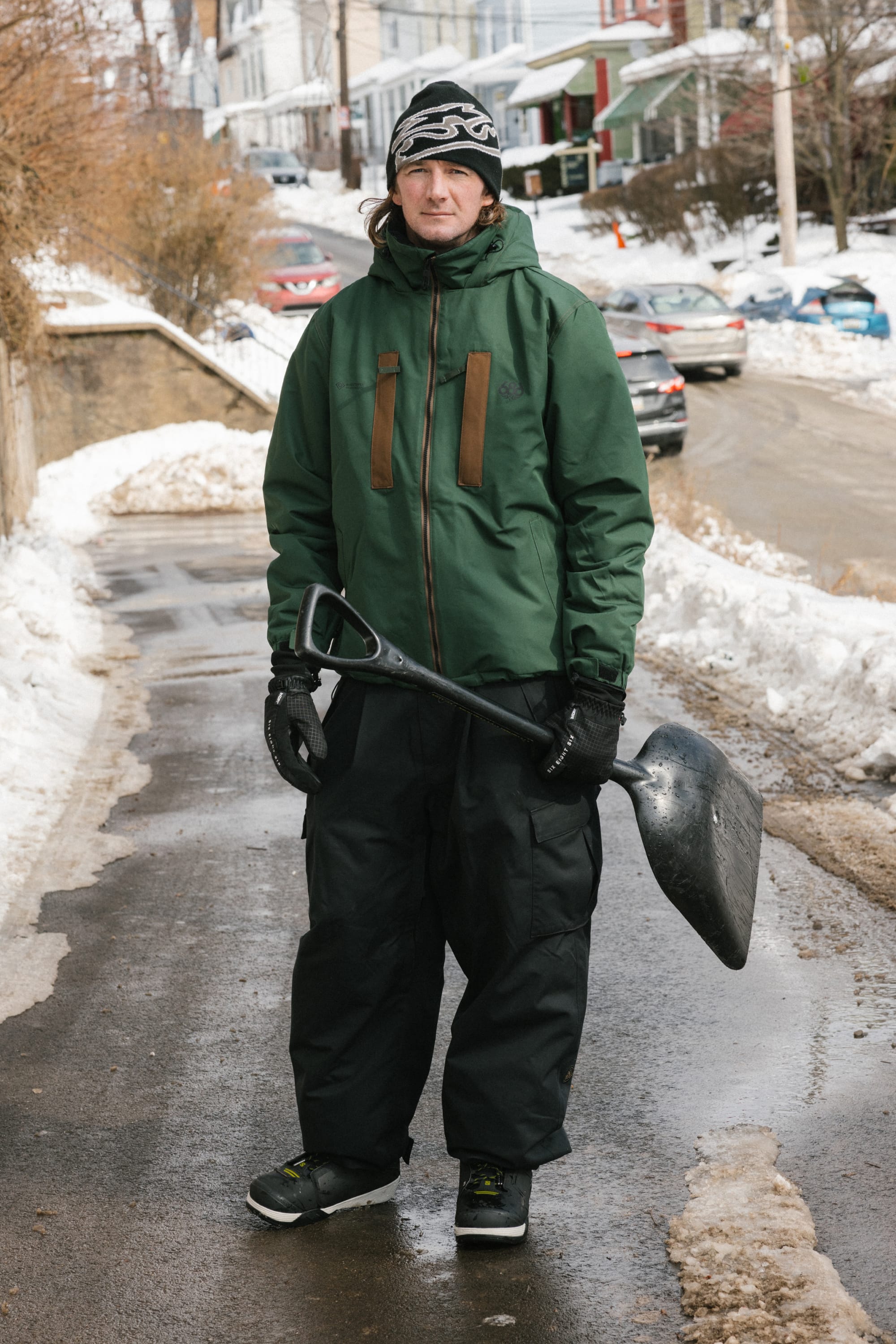 A person in winter clothing stands on a snowy street holding a large snow shovel, bundled up in the 686 Men’s WINDSTOPPER® Guideline Insulated Jacket by GORE-TEX® LABS. Houses and parked cars line the background.