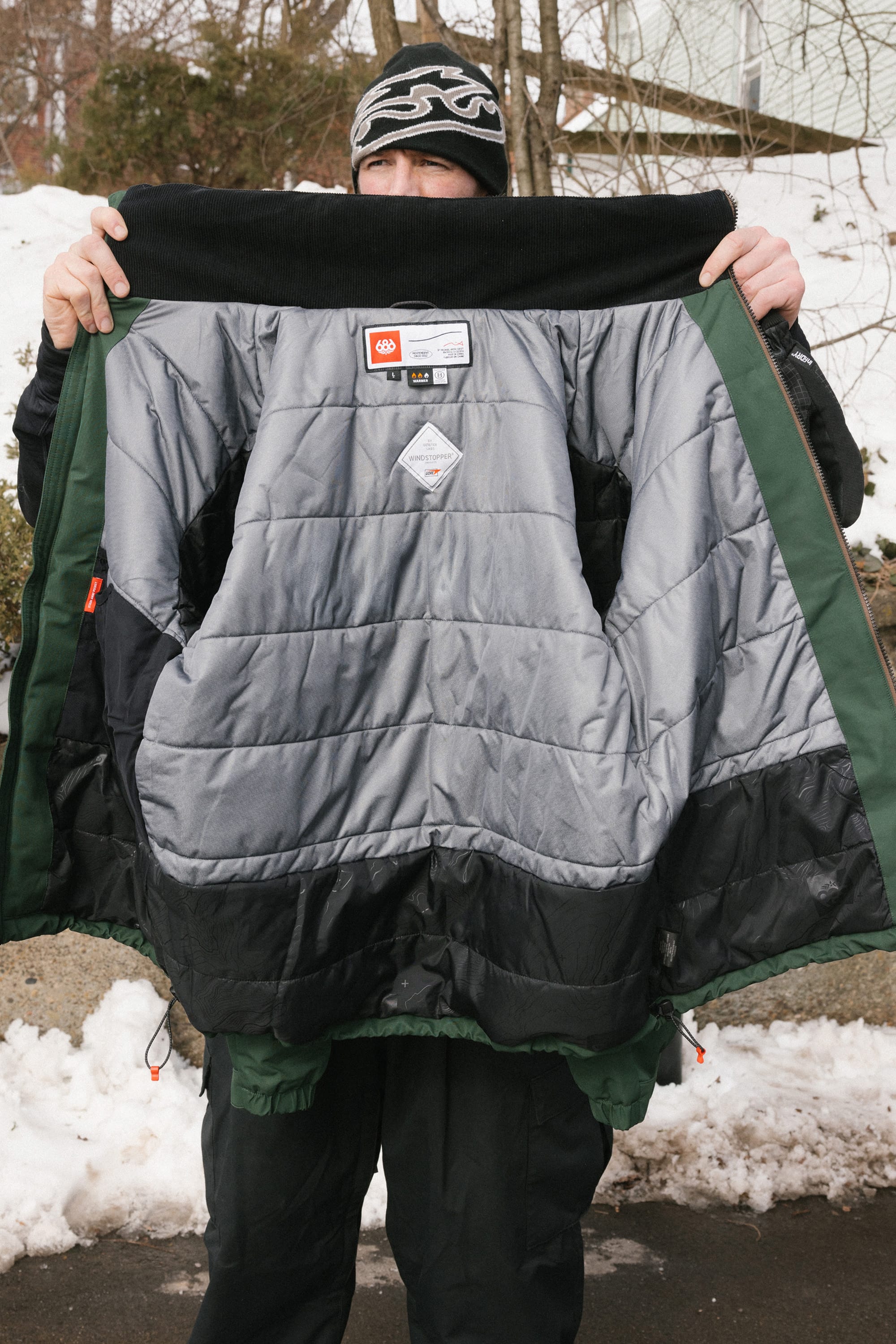A person holds open the 686 Men’s WINDSTOPPER® Guideline Insulated Jacket by GORE-TEX® LABS outdoors in winter, displaying its quilted interior and labels, with snow and trees in the background.
