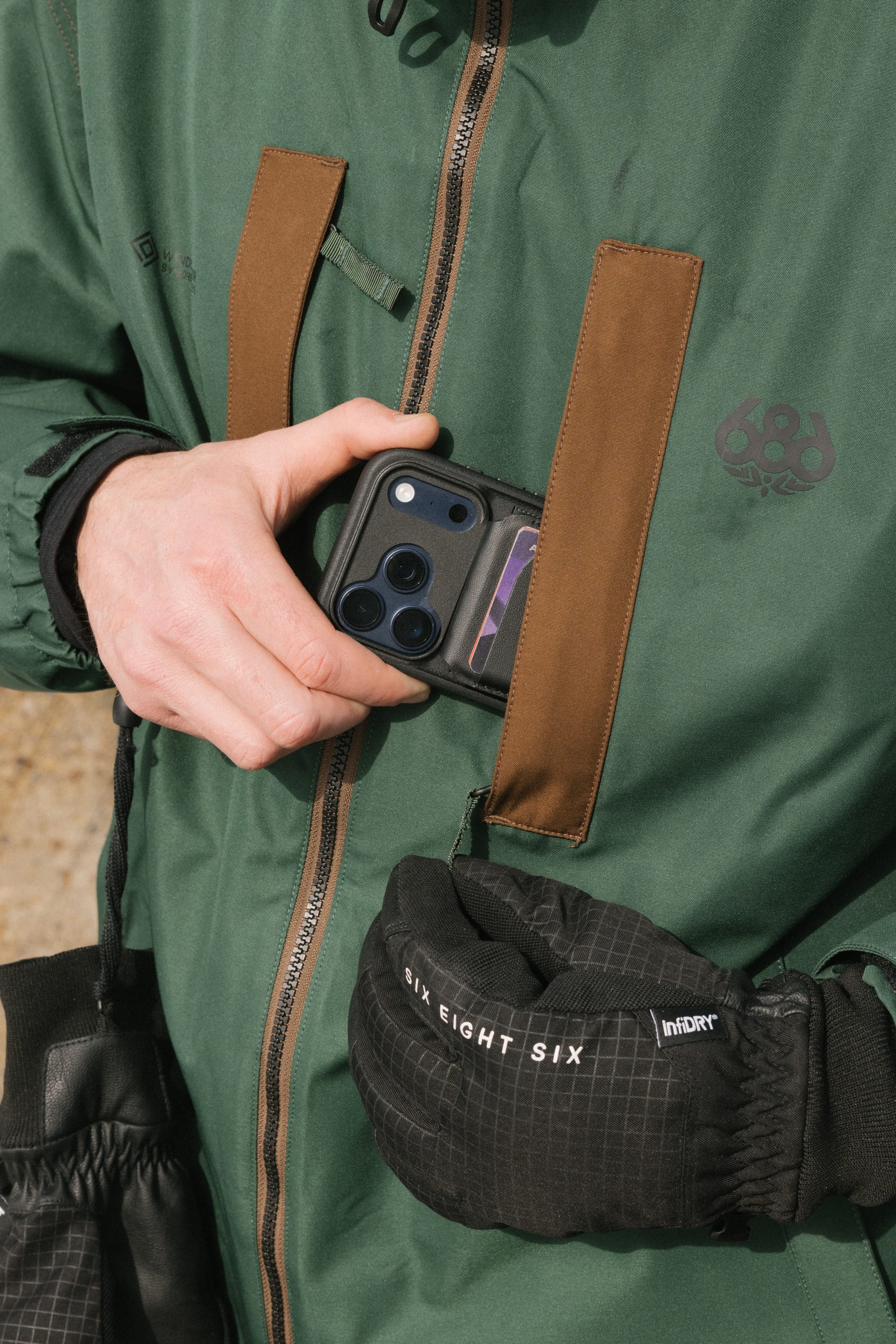 A person wearing the 686 Men’s WINDSTOPPER® Guideline Insulated Jacket by GORE-TEX® LABS in green slips a triple-camera smartphone into a brown chest pocket, while carrying a black pouch labeled 686.
