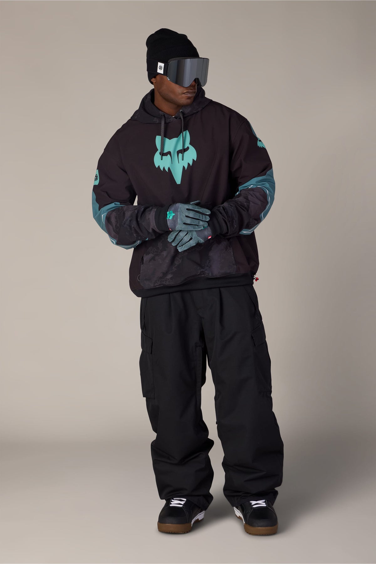 A person wearing a black beanie, mirrored ski goggles, a 686 Men’s Fox Racing Waterproof Team Hoody, black pants, and patterned gloves standing against a light grey background.