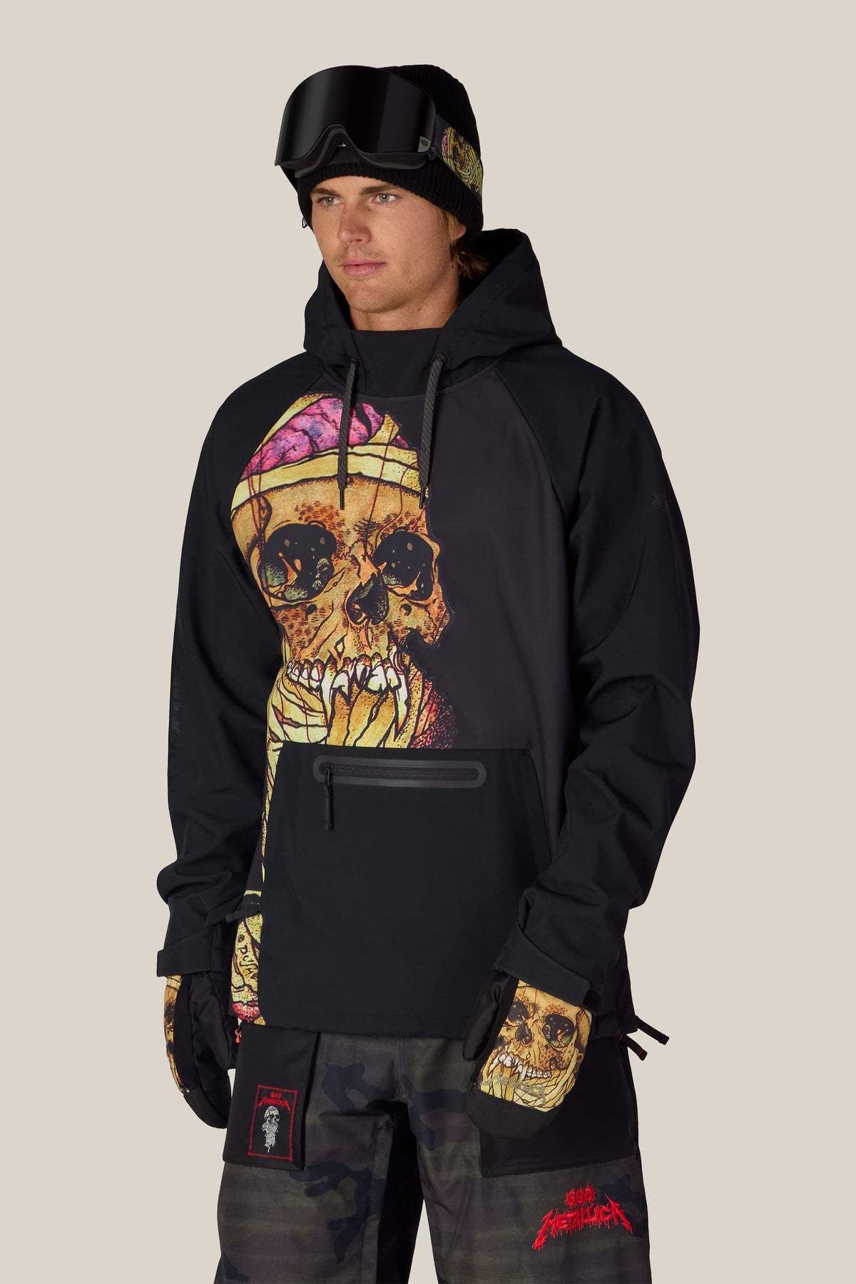 A person wearing a 686 Men’s Metallica Waterproof Hoody with a large skull graphic on the chest, a black beanie with goggles, and camouflage pants.