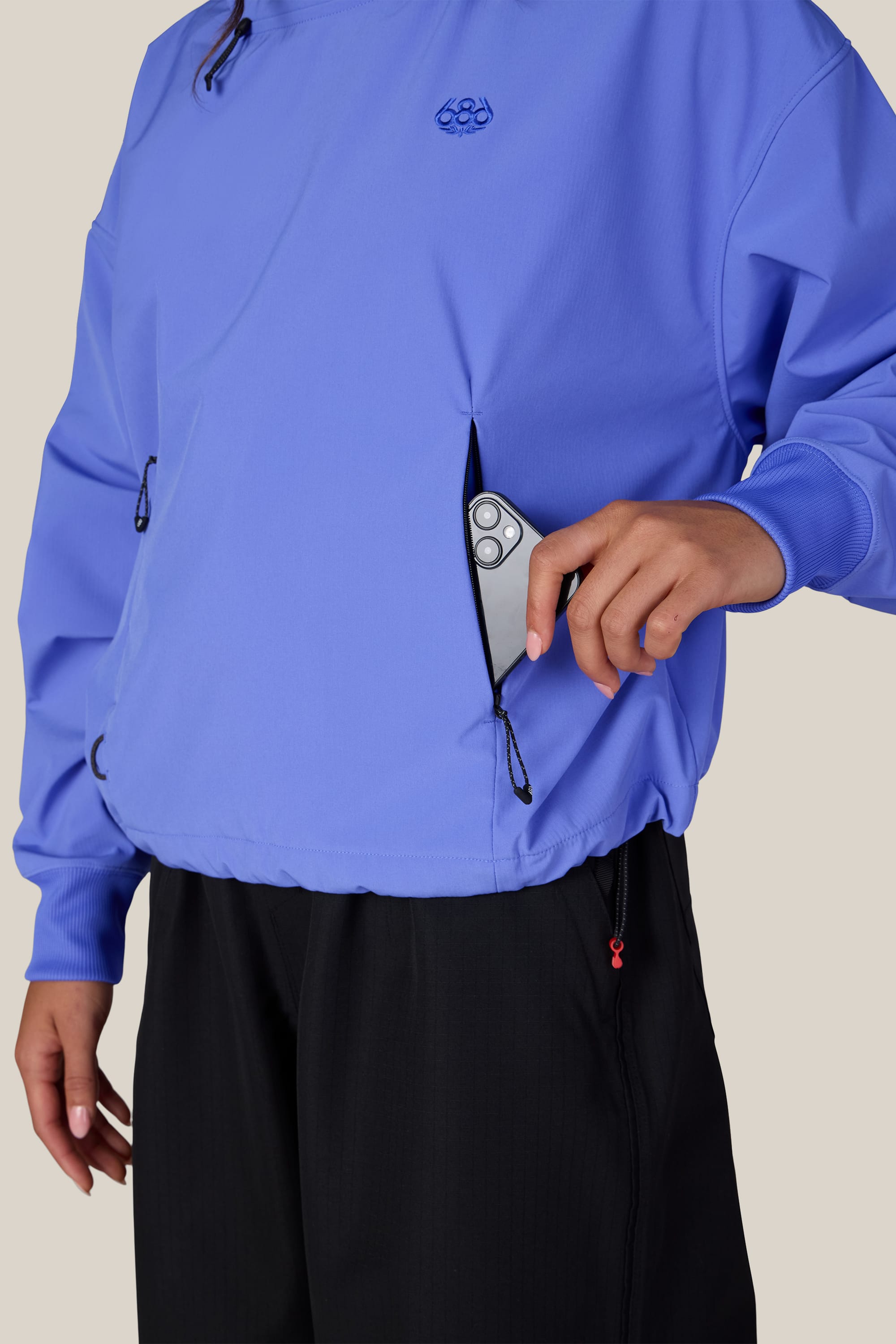 Wearing the 686 Women’s Waterproof Team Crop Hoody in blue and black pants, a person puts a silver smartphone into the zippered front pocket of the sweatshirt.