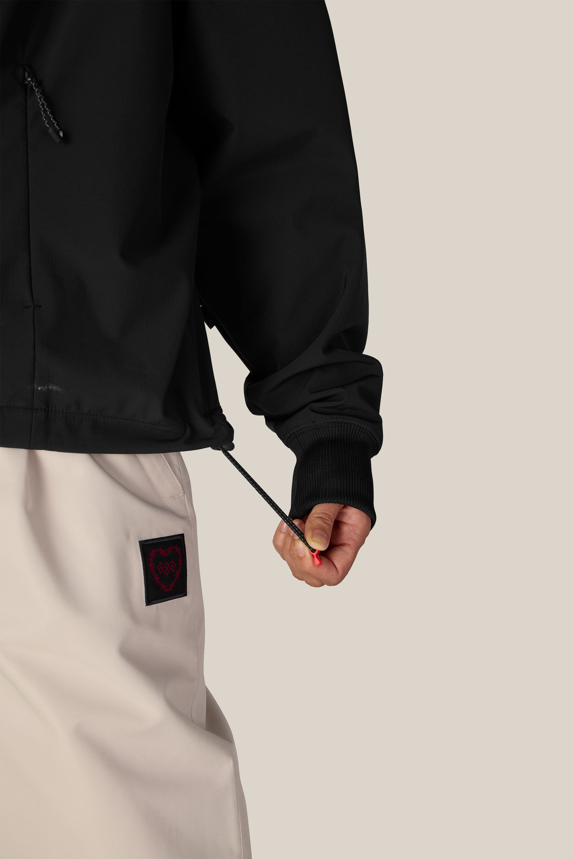 A person wearing the 686 Women’s Waterproof Team Crop Hoody in black tightens the jacket’s drawstring. They are also wearing light beige pants with a black patch featuring a red heart on the pocket. The background is solid beige.