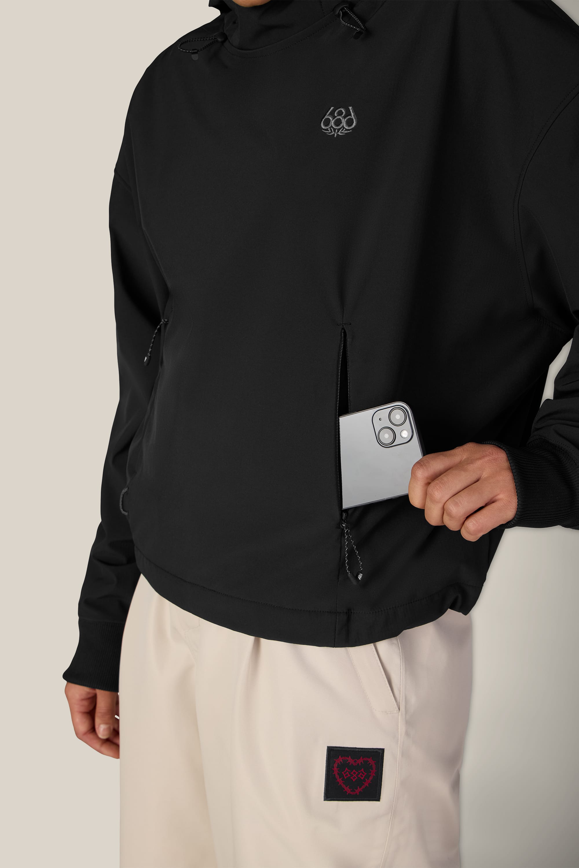 A person wearing the 686 Women’s Waterproof Team Crop Hoody in black and light beige pants slides a silver smartphone into the hoody’s zippered pocket, with a small patch featuring a red heart design visible on the pants.