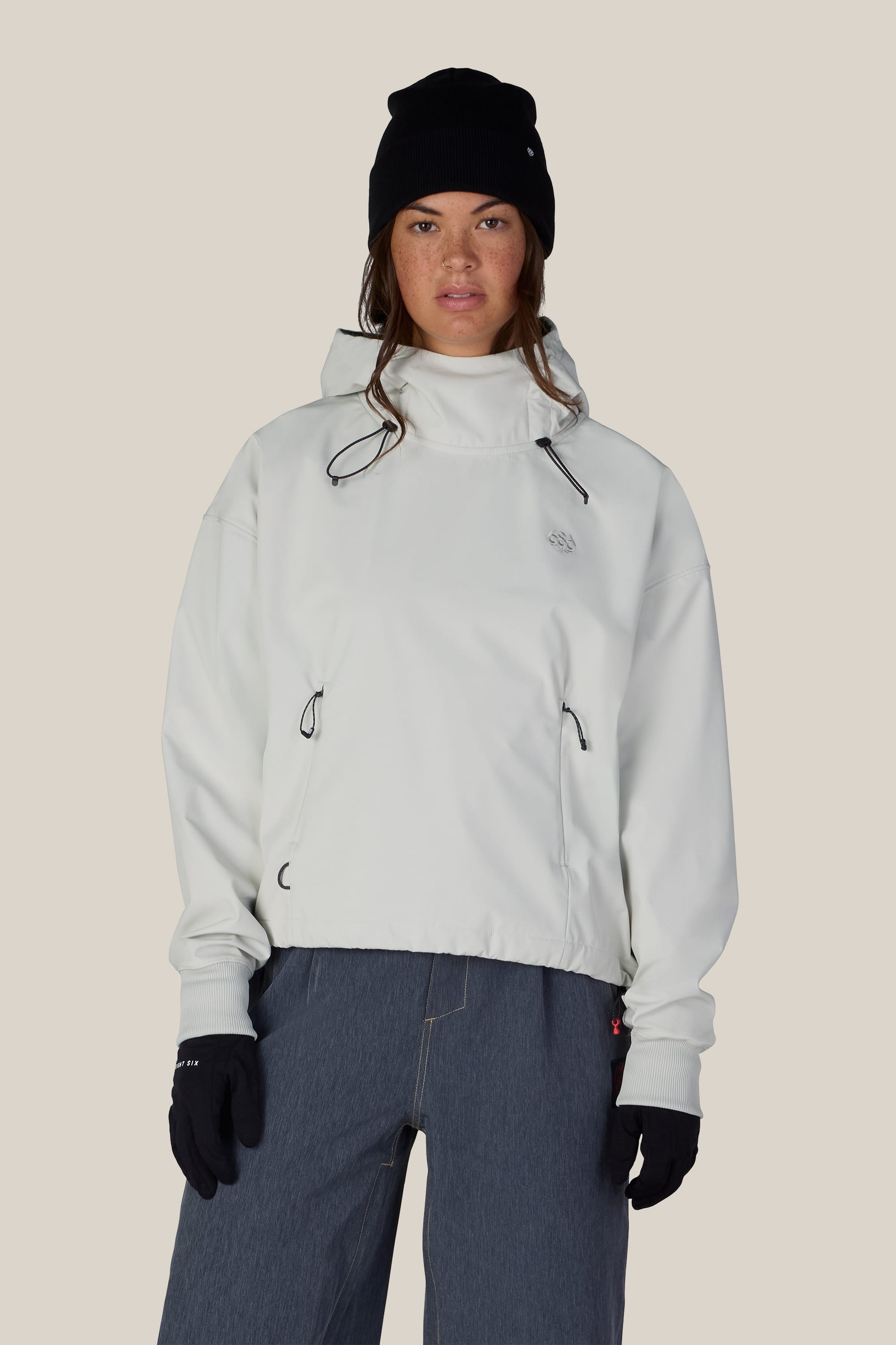 A person wearing the 686 Women’s Waterproof Team Crop Hoody with a white hood, along with a black beanie, black gloves, and blue pants stands facing forward against a plain beige background.
