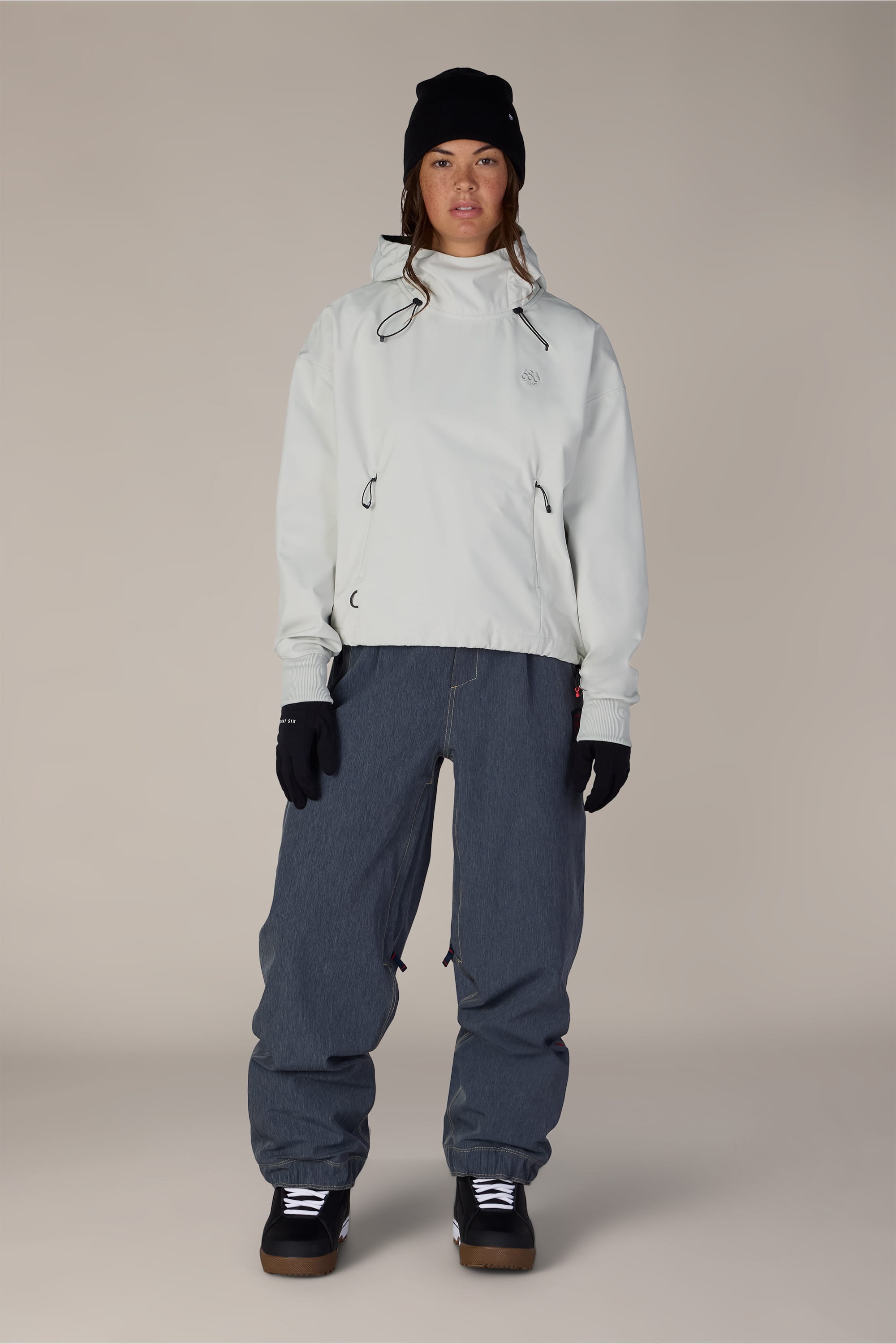 A person wearing the 686 Women’s Waterproof Team Crop Hoody in white, along with dark baggy pants, black gloves, a black beanie, and black sneakers, stands against a plain light background looking directly at the camera.