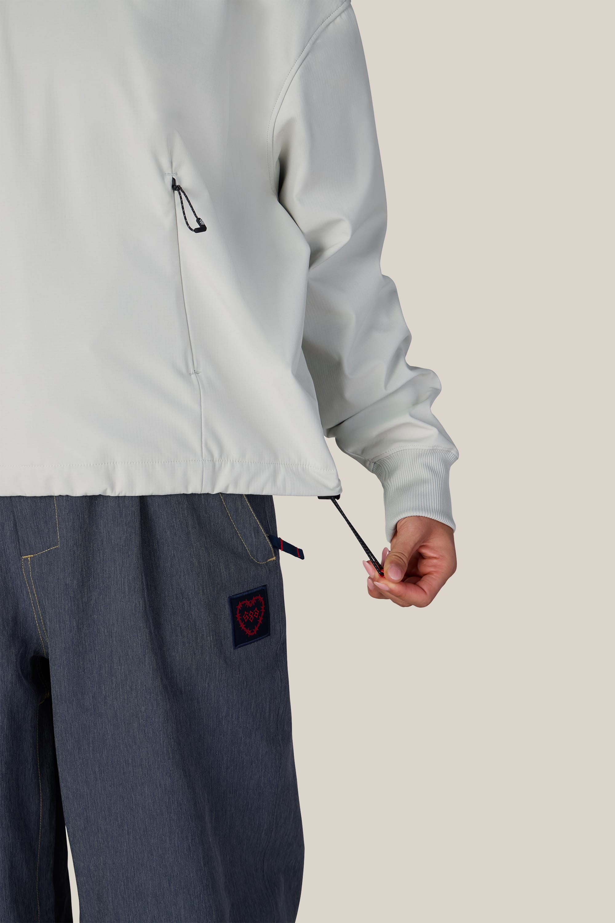 A person adjusts the drawstring on the hem of a white 686 Women’s Waterproof Team Crop Hoody, paired with loose blue pants featuring a small heart patch on the pocket, set against a plain beige background.