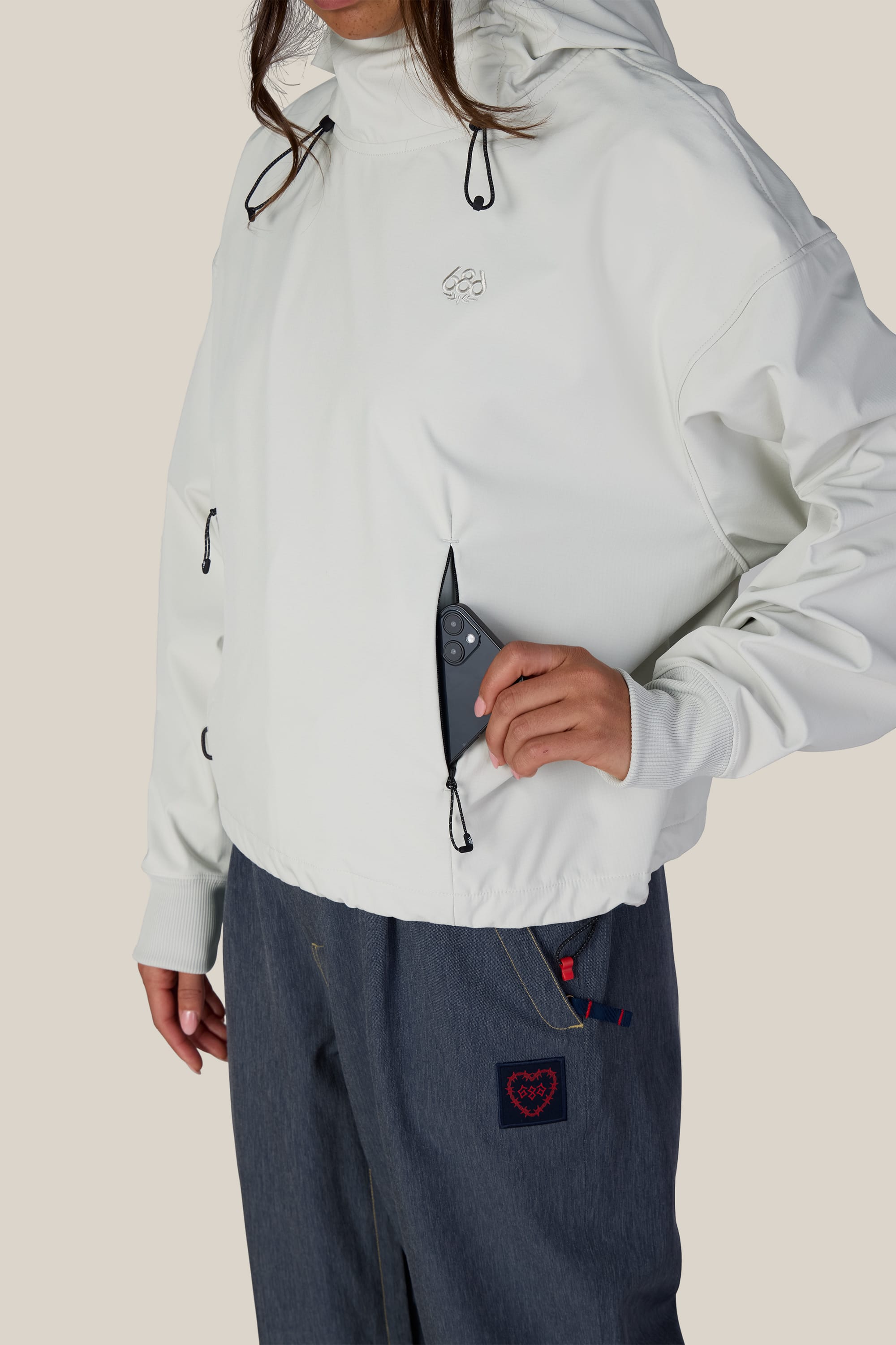 A person wearing the 686 Women’s Waterproof Team Crop Hoody in white, featuring black drawstrings and a zippered chest pocket for stashing a smartphone, pairs it with blue pants accented by a red embroidered heart patch.
