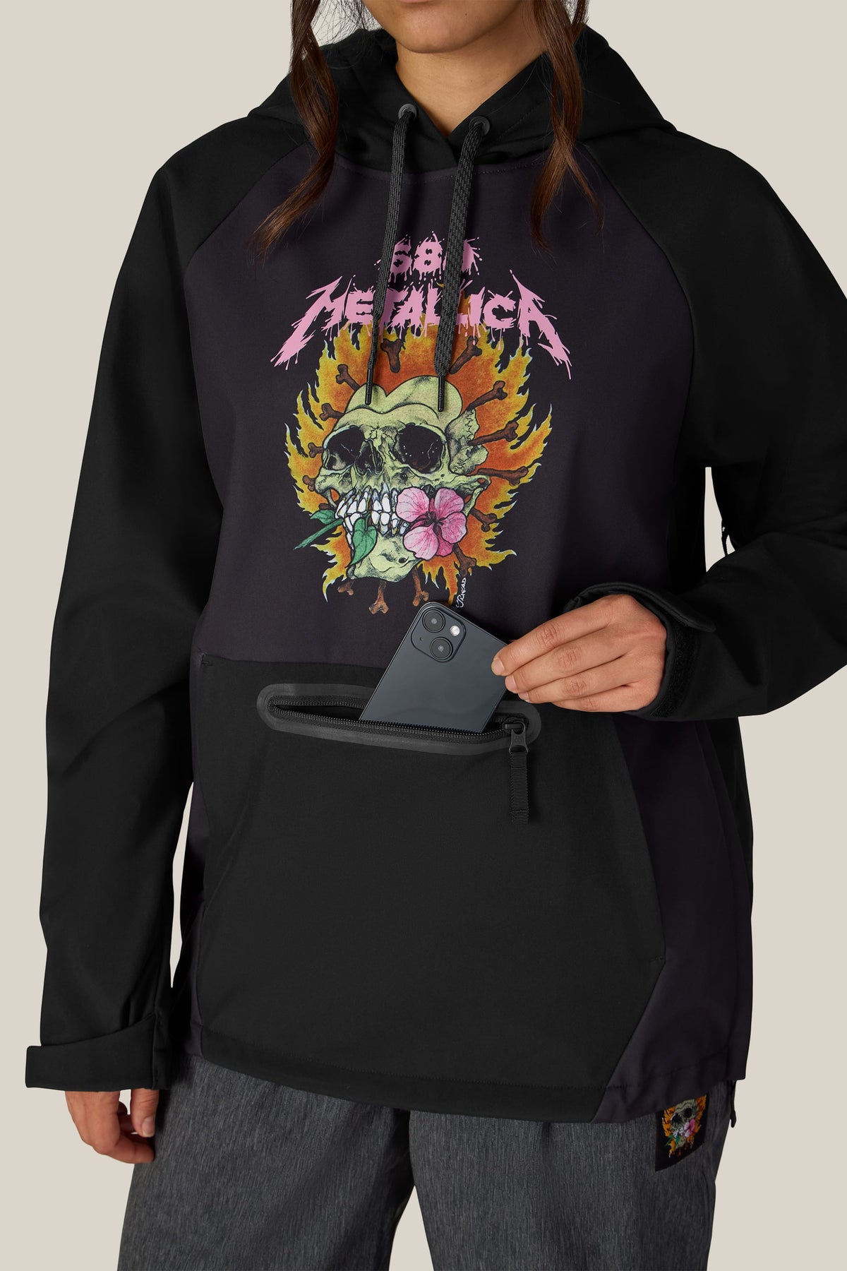 A black waterproof 686 Women’s Metallica Waterproof Hoody with a skull-in-flames graphic and a zippered chest pocket containing a smartphone.