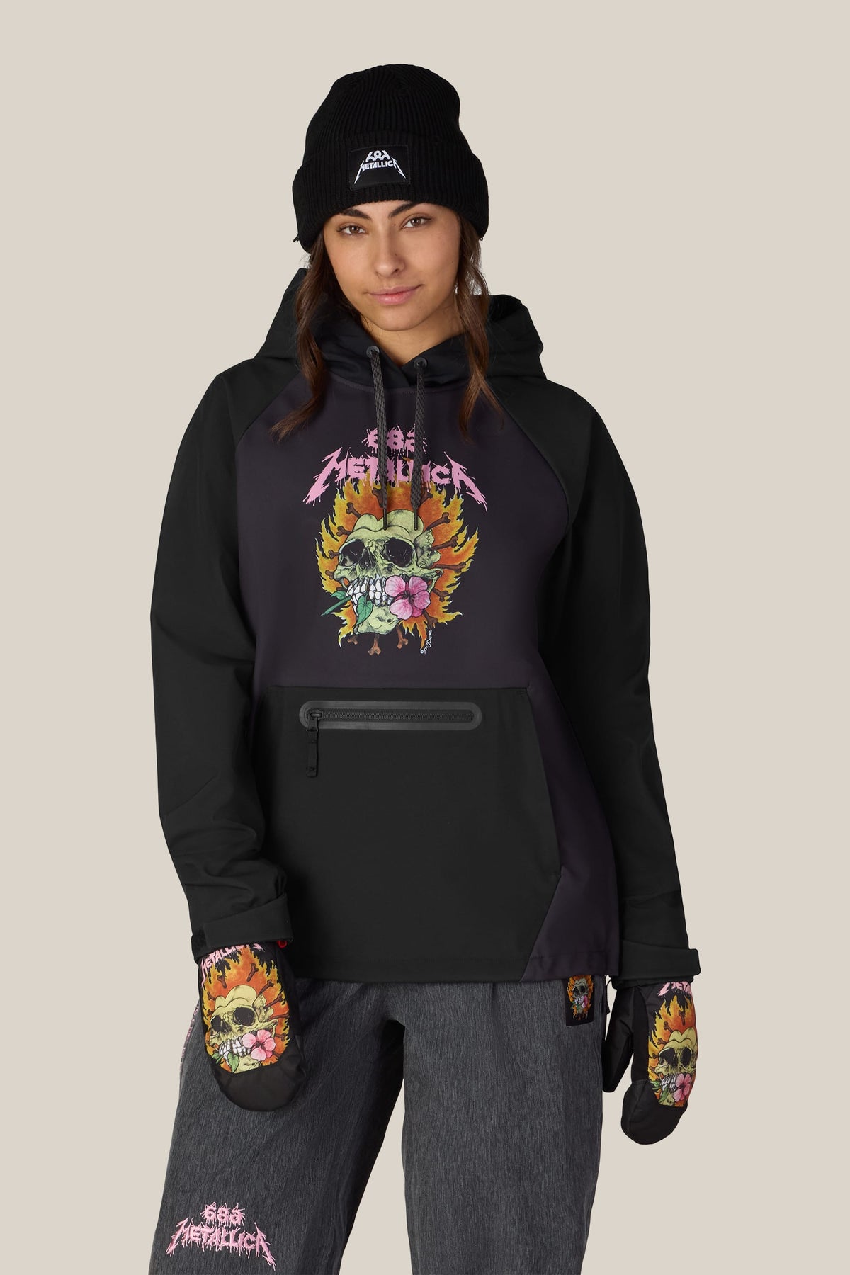 A person wearing a black 686 Women’s Metallica Waterproof Hoody and matching mittens, both featuring a graphic of a skull with flowers and flames.