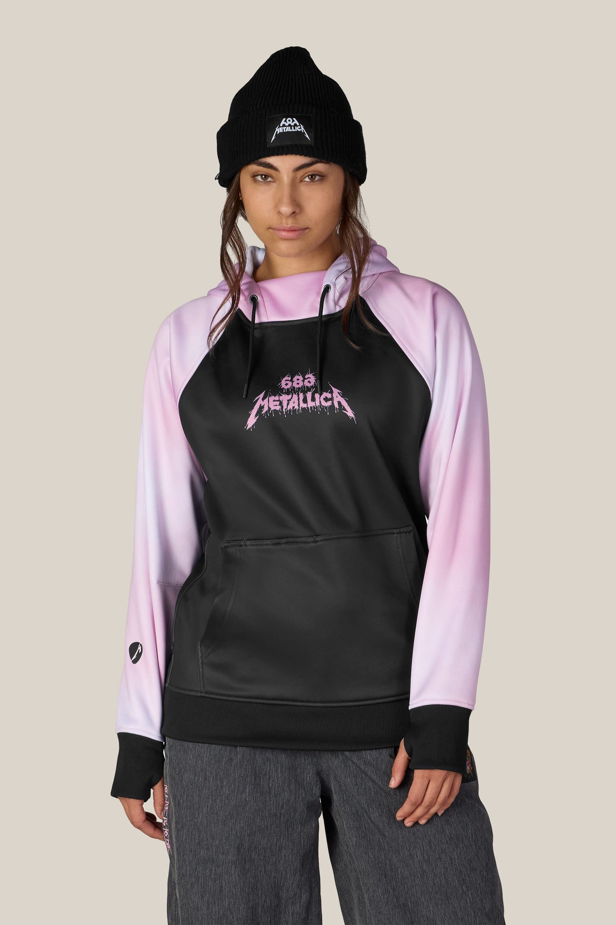 A person wearing a black knit beanie with a Metallica logo and a 686 Women’s Metallica Bonded Fleece Pullover Hoody featuring a pink Metallica graphic on the chest.