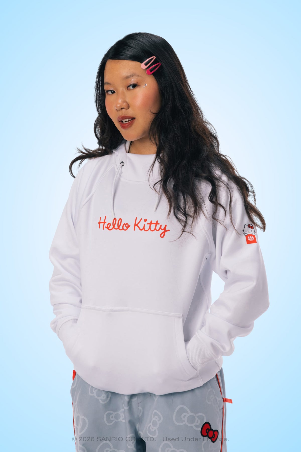 A woman with long dark hair stands against a light blue background, looking forward with her hands in her pockets. She is wearing the 686 Women’s Hello Kitty® Bonded Fleece Pullover Hoody by 686 and patterned gray pants. Her look is complemented by pink hair clips and natural makeup.