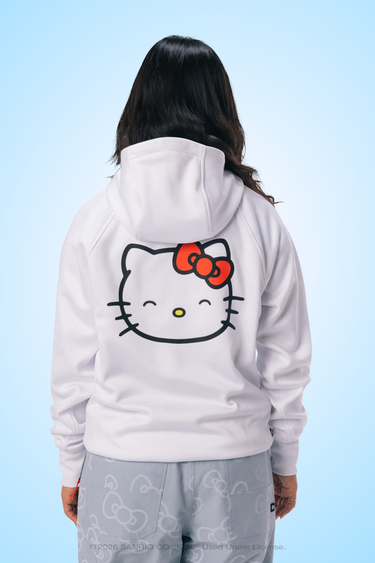 A person stands against a light blue background, facing away from the camera, wearing the 686 Women’s Hello Kitty® Bonded Fleece Pullover Hoody by 686, which features a large Hello Kitty face and red bow on the back and is made from soft polyester fleece material.