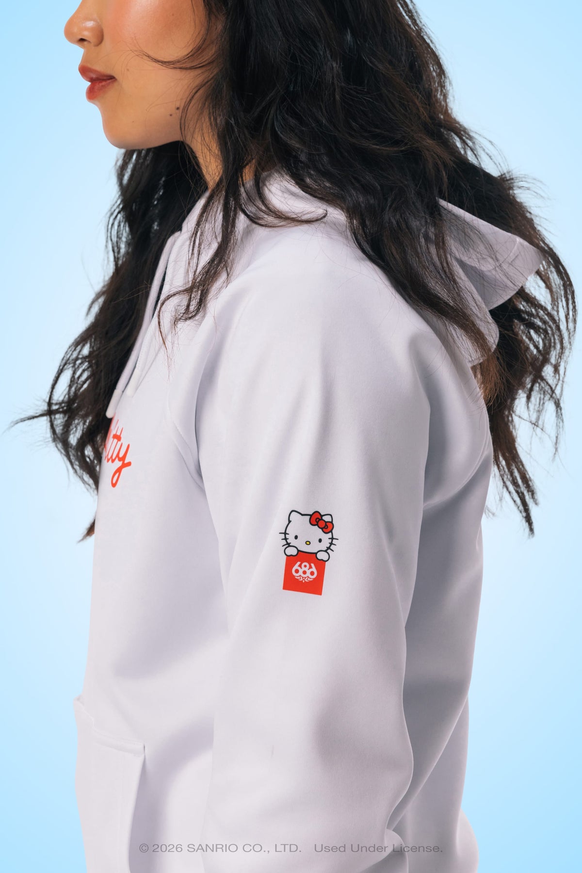 A woman with long dark hair wears the 686 Women’s Hello Kitty® Bonded Fleece Pullover Hoody—a white polyester fleece jacket by 686 featuring a small Hello Kitty graphic on the sleeve—set against a light blue background.