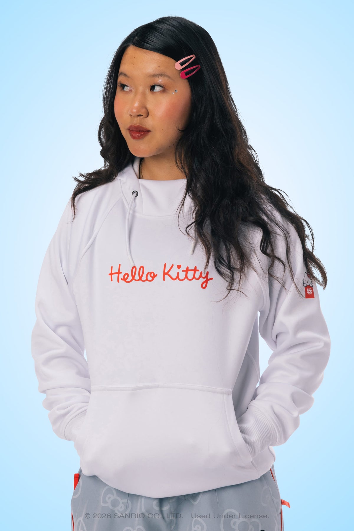 A woman with long dark hair, wearing pink hair clips and red lipstick, models the 686 Women’s Hello Kitty® Bonded Fleece Pullover Hoody. She stands against a light blue background, looking to the side with her hands in the front pocket.