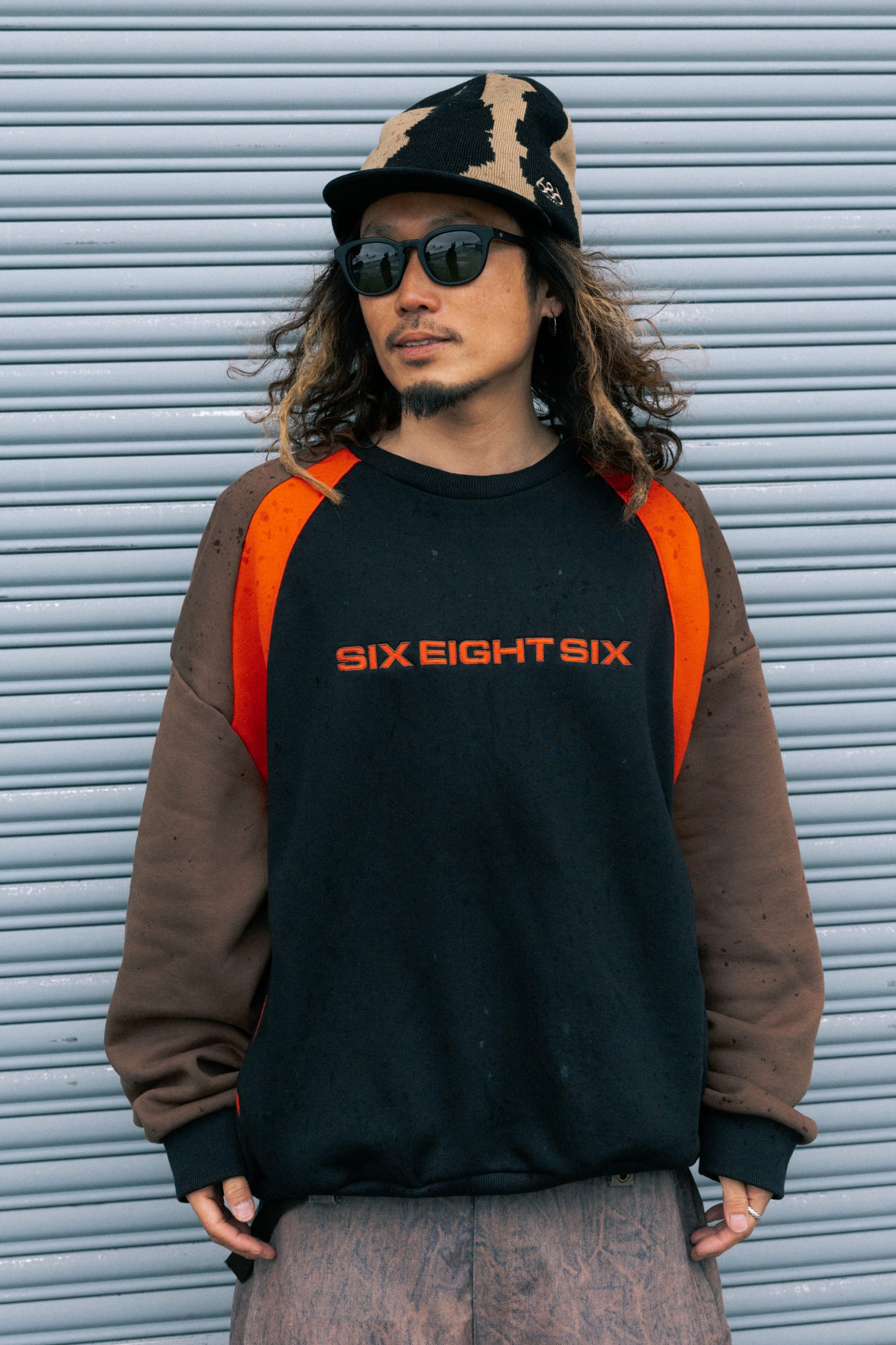 A man stands in front of a corrugated metal background, wearing sunglasses, a cap, and the 686 Reserved® Premium Heavyweight Crewneck—a black men’s sweatshirt with orange accents by 686.