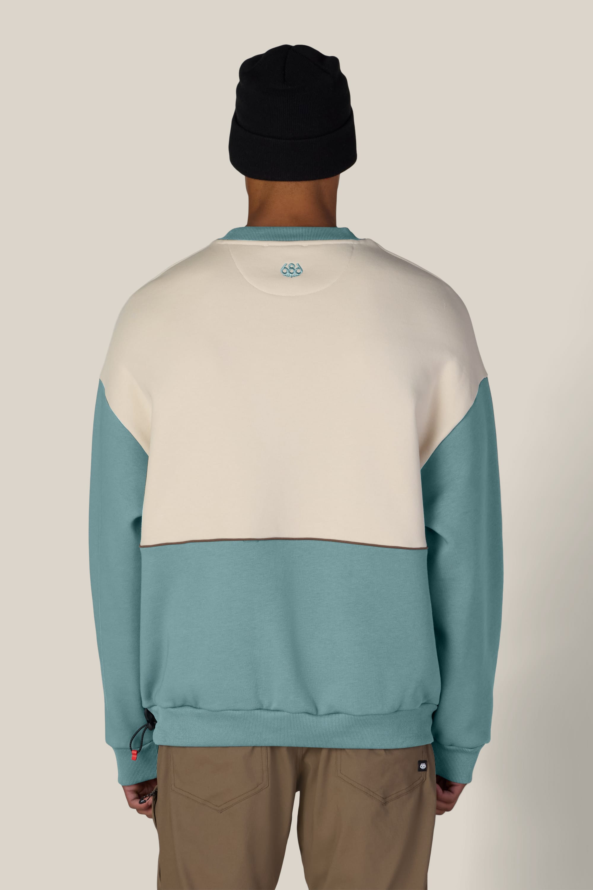 A person wearing the 686 Reserved® Premium Heavyweight Crewneck by 686 in a color-blocked cream and teal design, paired with tan pants and a black beanie, stands with their back to the camera against a plain background.