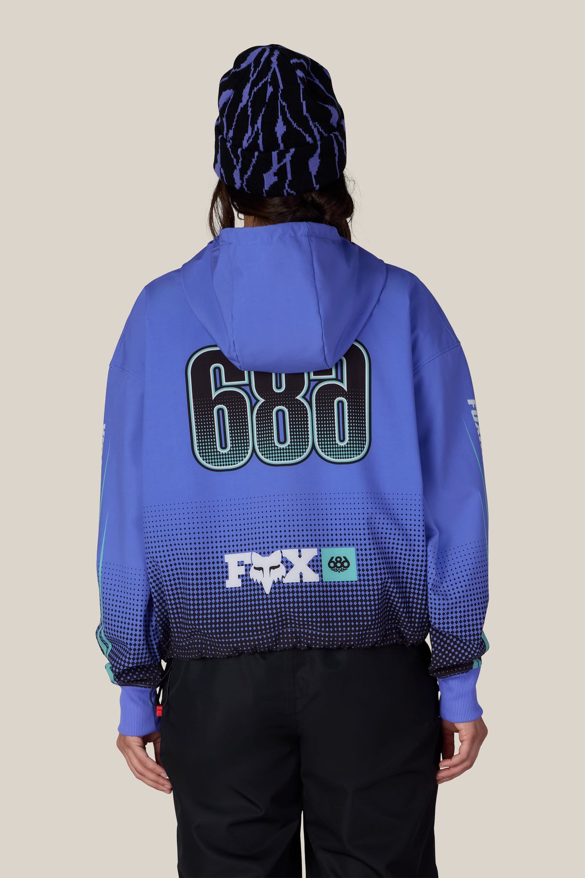 A person from behind wearing a 686 Women’s Fox Racing Waterproof Crop Hoody paired with a patterned blue and black beanie.