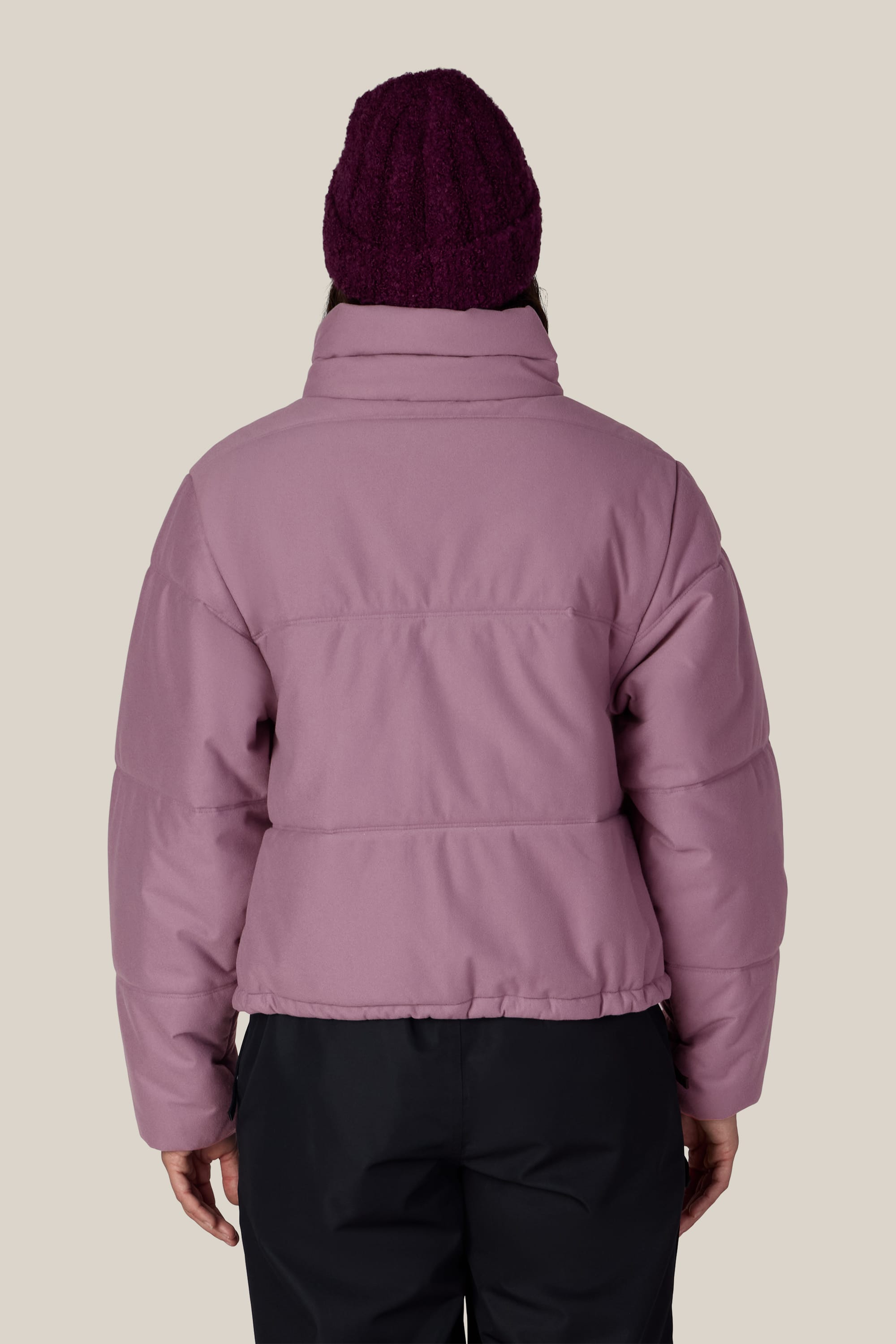 Person facing away, wearing the 686 Women’s Gracious Puffer Jacket in mauve, paired with black pants and a dark purple knit beanie, standing against a plain light background.