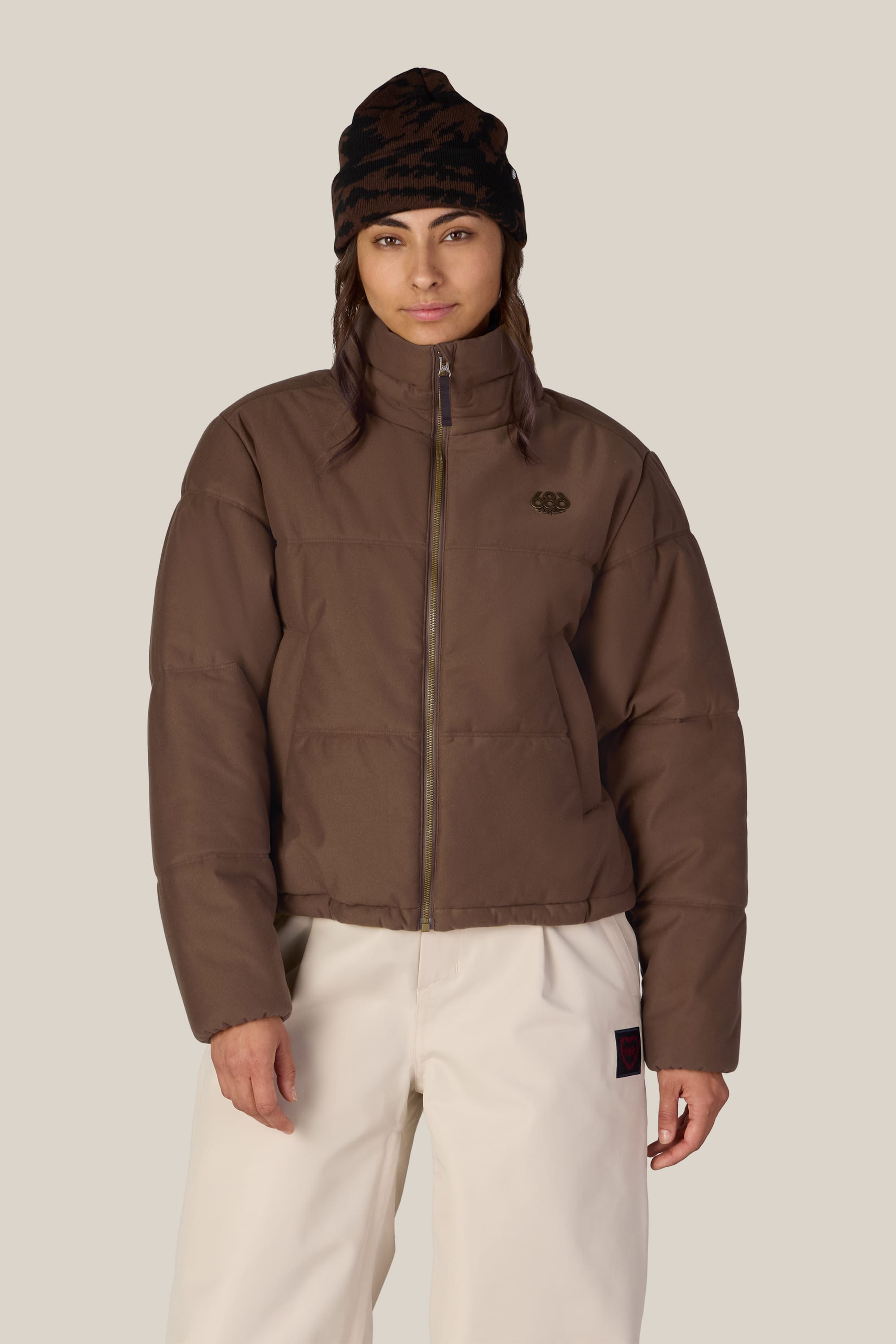 A person wearing the 686 Women’s Gracious Puffer Jacket in brown, paired with white pants and a dark knit beanie, stands facing forward against a plain light background.